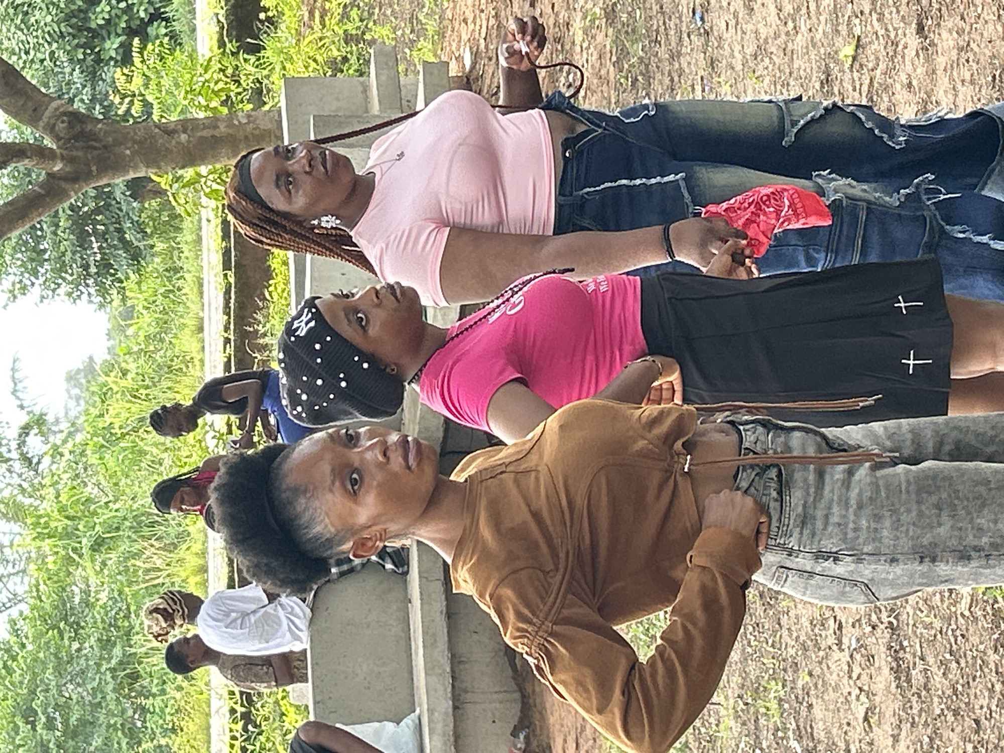 A group of people stand outdoors in Ihiagwa, Nigeria. The foreground features three women, one wearing a brown jacket and light-colored jeans, another in a pink t-shirt and black skirt with white cross designs, and a third in a light pink t-shirt and ripped jeans with a red bandana. Additional people stand further back, with some sitting on a low concrete structure. Trees and vegetation are visible in the background. The ground appears to be dirt and grass.