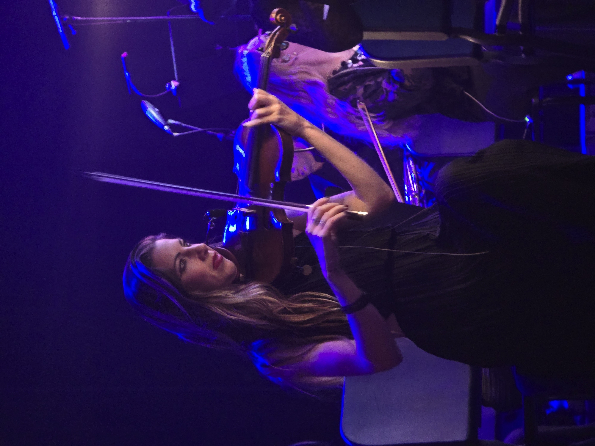 A woman plays the violin on a dimly lit stage, with purple and blue spotlights casting shadows and highlights. She is wearing a dark, sleeveless dress and has long, wavy hair. The violin is a dark wood color, and she holds the bow with her right hand. Her left hand is positioned on the fingerboard. In the background, another person with long hair and a patterned top is visible, also appearing to be part of a performance. Several microphones on stands are positioned around the stage. The overall impression is of a musical performance in a concert venue. The image does not provide specific information to determine the location in the city of Los Angeles, United States of America.