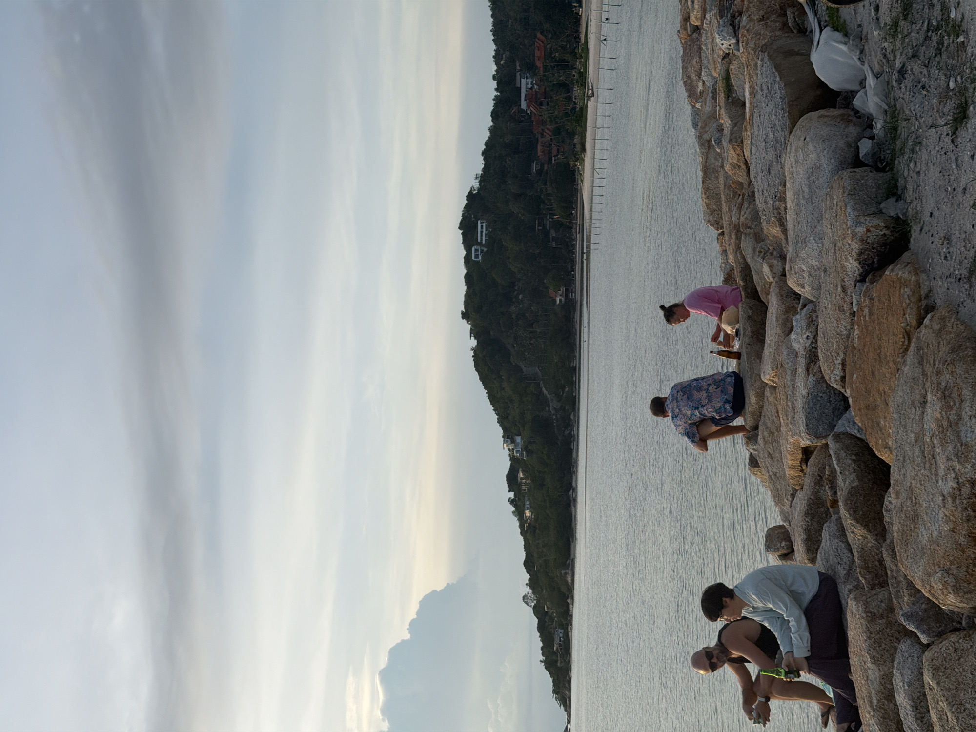 In ตำบลเกาะพะงัน, Thailand, on a rocky shoreline, four people are seated near the water's edge. On the left, two men sit on large rocks, one holding a bottle. To their right, a person in a light blue shirt and dark pants is seated with a bottle of beer in hand. Further to the right, a person in a bright pink shirt is also seated on the rocks. The sea is calm with gentle ripples, and a verdant, hilly coastline with scattered houses is visible in the background under a sky with scattered clouds. A line of wooden posts extends into the water in the middle ground.