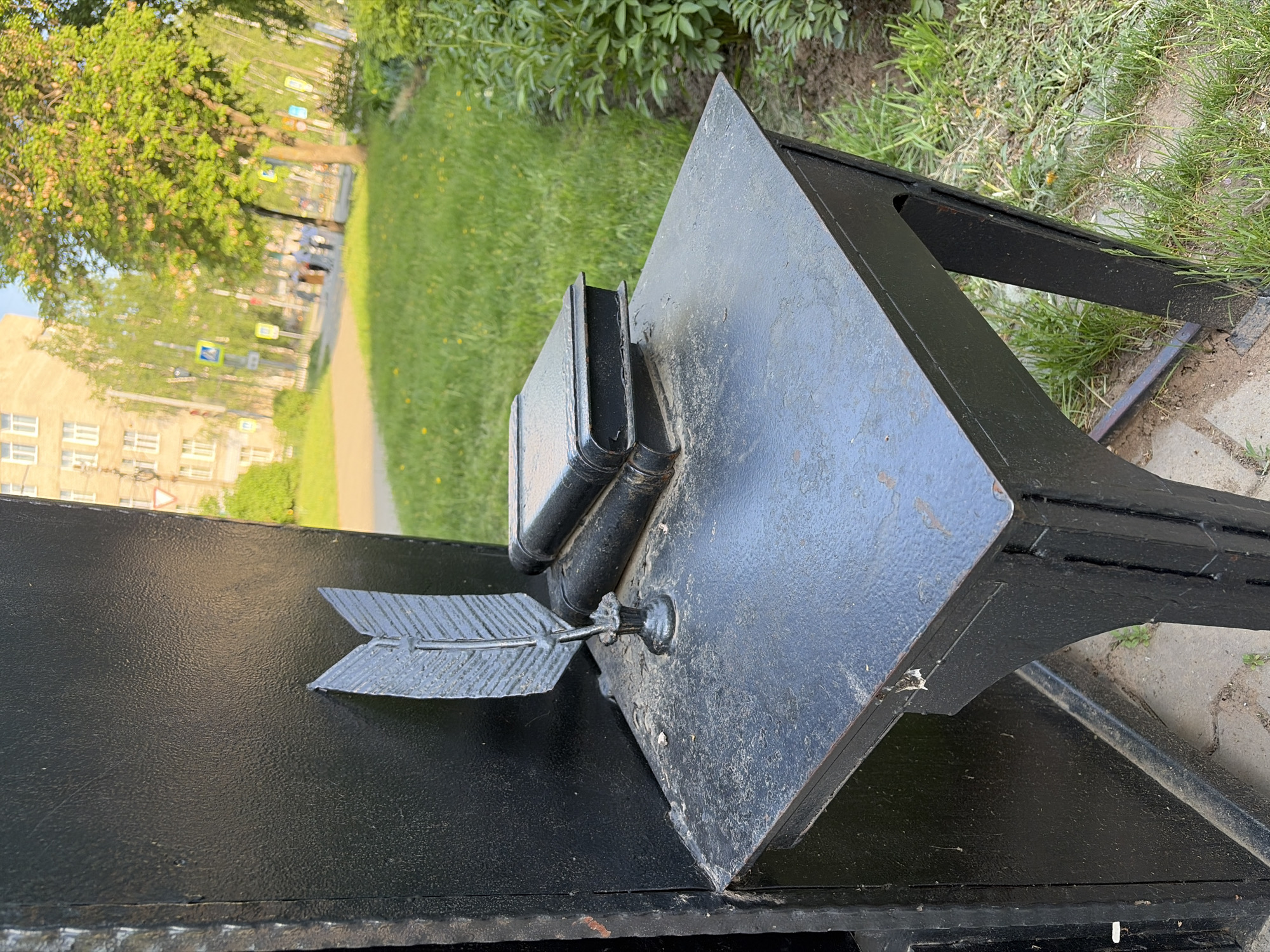 The image shows a metal monument in Chaikovsky, Russia. The monument consists of a black metal structure resembling a desk or lectern, with an open book and a pen represented by a metal arrow attached to the side. The surrounding environment includes a green lawn, trees, a sidewalk, and a building in the distance.