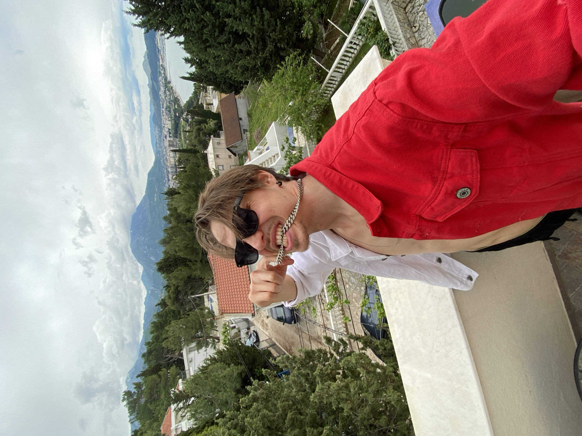 A man, likely in his 20s or 30s, is captured in a selfie on a balcony, presenting a playful or confident expression. He is wearing sunglasses, a white collared shirt, and a red denim jacket, and holds a chunky silver-colored chain necklace up to his mouth with a slight grin. His hair is light brown and somewhat tousled.
The setting is a scenic outdoor location, likely a terrace or balcony, offering an expansive view. In the background, a coastal town with numerous white buildings and reddish-brown tiled roofs spreads across a hilly landscape, interspersed with lush green trees, particularly cypress-like evergreens. Beyond the town, majestic mountains rise, and in the distance, a calm body of water, presumably the sea, meets the horizon under a cloudy sky. The weather appears overcast but bright, indicating daytime. The scene strongly suggests a Mediterranean or Adriatic coastal environment, consistent with the location context of Šušanj, Montenegro. No visible text is present in the image.