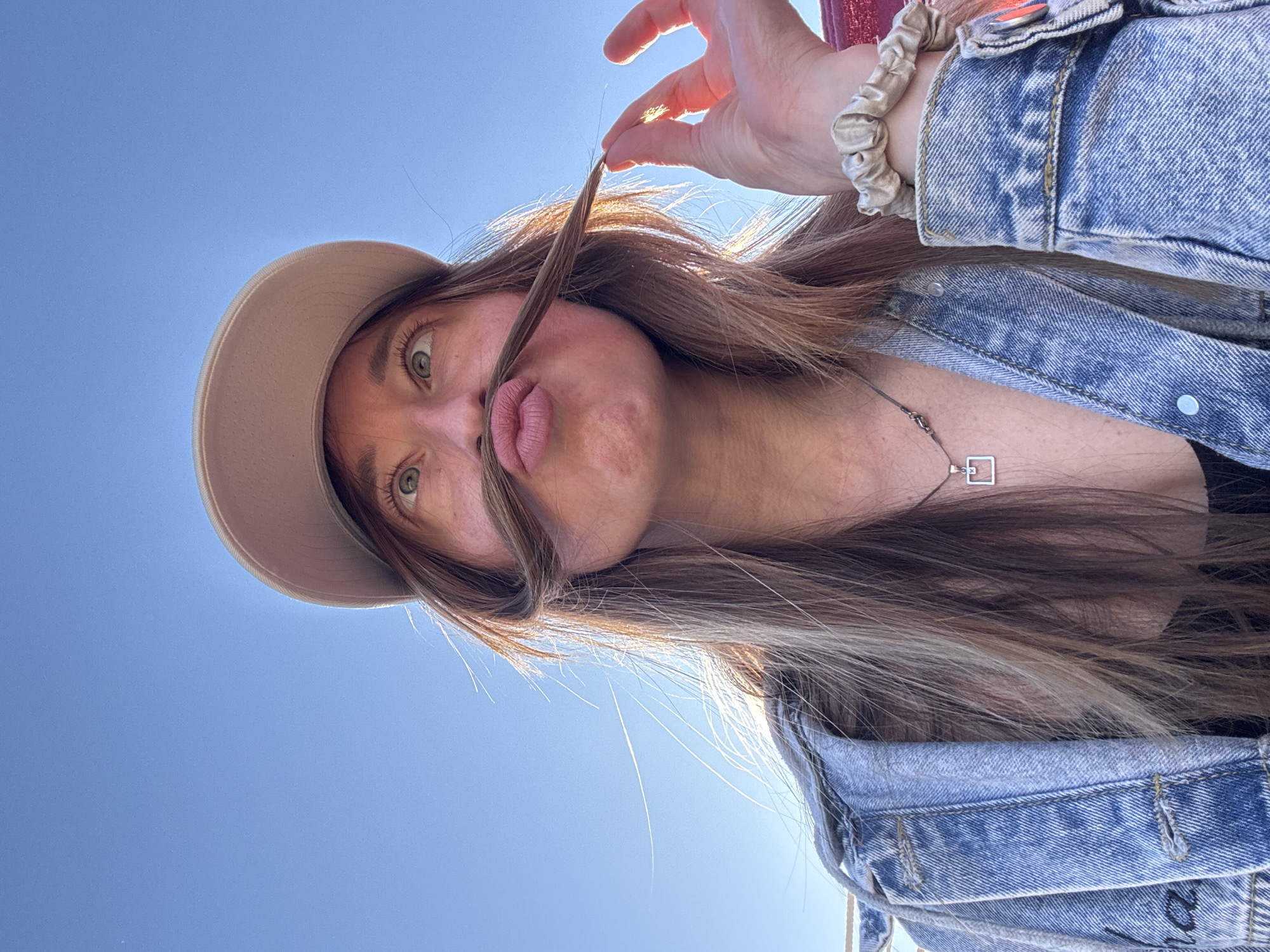 A woman wearing a light tan hat and a blue denim jacket is photographed from a low angle, looking up into the camera against a clear blue sky. She is holding a strand of her brown hair under her nose, forming a makeshift mustache. She has green eyes and pursed lips. A silver necklace with a square pendant is visible. A scrunchie is on her wrist. The photograph appears to be taken outdoors. There is no evidence to indicate the location is Прикамский, Russia.