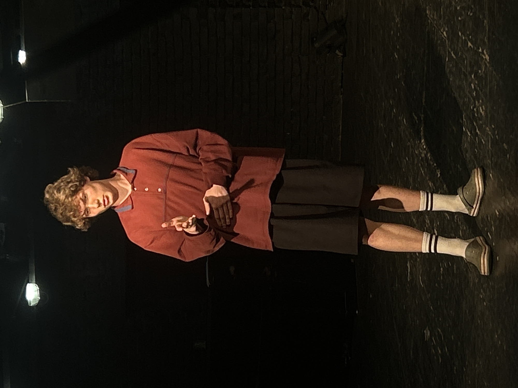 A young individual stands on what appears to be a dimly lit stage or performance space. The person, with light brown curly hair, is positioned centrally in the frame, looking slightly towards the left with a serious or contemplative expression. They are wearing a long-sleeved collared shirt in a muted red or rust color, dark-colored shorts, white athletic socks with two black stripes near the top, and grey sneakers with white soles.
Their hands are raised to chest height, with the left hand resting on the right forearm, and the right hand extended with fingers slightly apart, as if mid-gesture during a speech or performance. The background is predominantly dark, suggesting a deep stage or room, with a dark, textured wall behind the subject. The floor is also dark and appears to be made of wood, reflecting some of the light.
Several bright light sources are visible in the upper left background, indicating artificial stage lighting that illuminates the subject primarily from the left side, casting some shadows. The overall setting implies an indoor environment, likely a theatre, studio, or rehearsal room. No visible text is present in the image.