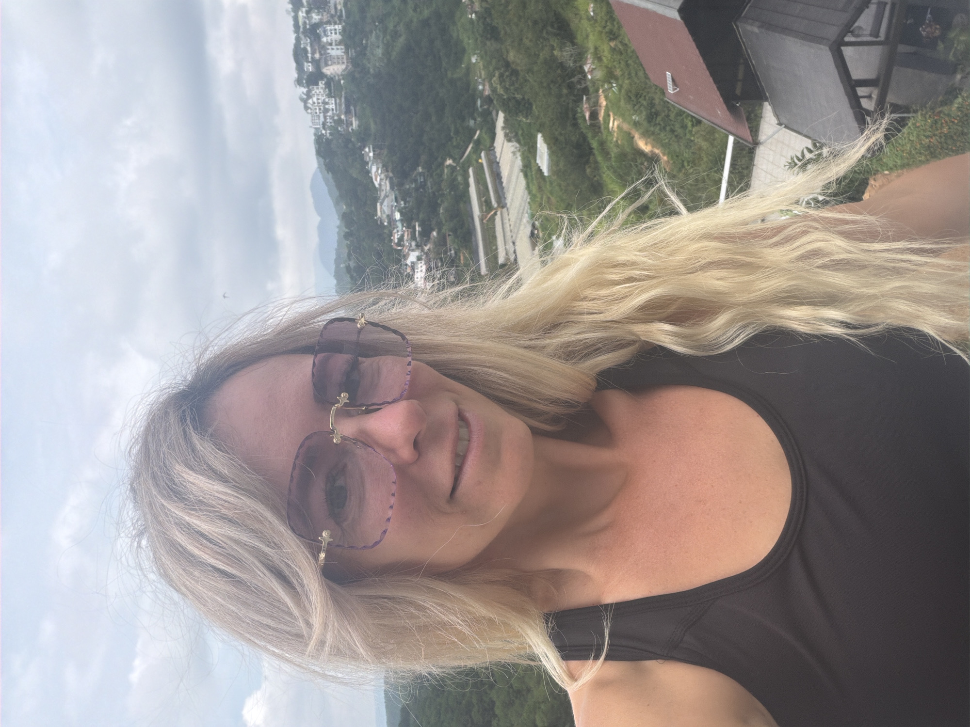 A blonde woman with light skin and blue eyes is taking a selfie outdoors. She is wearing large, rectangular sunglasses with a pink tint and gold frames, and a black tank top. Her blonde hair is long and wavy, falling over her shoulders. The background shows a hilly landscape with buildings scattered across the slopes. In the foreground, to the right of the woman, part of a dark-colored, angled roof structure is visible. The scene appears to be a scenic viewpoint, potentially overlooking a residential area or resort in Da Lat, Vietnam, given the architecture and lush greenery. The sky is overcast.