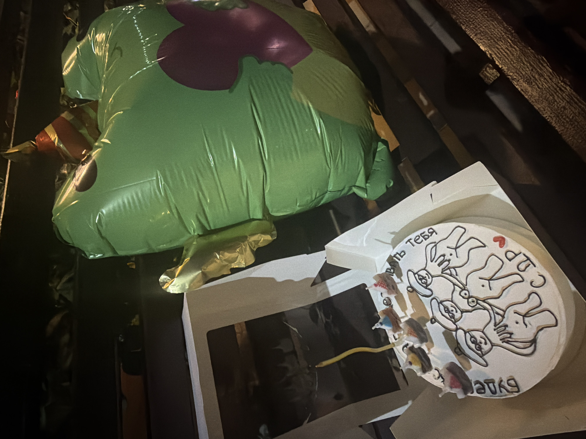 The image depicts a celebratory arrangement consisting of a cake and a large inflatable balloon, resting on a dark, textured wooden surface, possibly a bench, in a dimly lit environment, consistent with nighttime. The scene is situated in Anapa, Russia, as per the provided context.
In the foreground, slightly to the left, a white round cake is presented within an open cardboard cake box. The cake is frosted white and decorated with black icing outlines. Atop the cake, a single long, thin, wavy yellow candle stands upright, and six smaller, multi-colored (red, blue, yellow, brown, white) candles shaped like miniature shoes or boats are arranged around the cake's perimeter. The main decoration on the cake's surface features a drawing of three stylized, anthropomorphic figures. These figures wear hats resembling sailor caps or chef's hats, spectacles, and are depicted with their lower bodies exposed. They appear to be interacting in a playful or suggestive manner. Black icing text on the cake reads "Будем тебя С ДР" (roughly translating to "To your Happy Birthday") and a small red heart is drawn near "С ДР".
To the right of the cake, a large, inflated, shiny green balloon is visible. The balloon has an irregular, somewhat heart-like shape with a prominent darker purple-red heart graphic on its right side. Perched on top of the green balloon is a small, red, orange, and yellow striped conical party hat. A crumpled, reflective gold-colored item, possibly another smaller balloon or decorative element, is also attached near the top of the green balloon.
The background is dark and out of focus, showing indistinct shapes and varying shades of black and brown, suggesting additional wooden structures or ambient surroundings. No individuals are visible in the image; the focus is solely on the celebratory items.