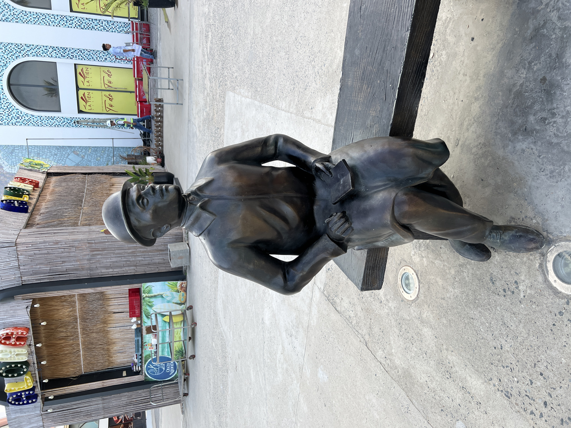 A bronze statue of a man in a hat and trench coat is seated on a dark wooden bench in a paved outdoor area. The statue is positioned in the foreground, facing slightly to the right, with his left hand resting on a book or notepad on his lap. Behind the statue, to the left, is a stall with bamboo walls and a colorful sign overhead that reads "GLOW". Various items are visible within the stall, including carts, crates, and decorative imagery. To the right of the statue, further in the background, is a modern building with large arched windows and blue and white mosaic tiling. Yellow signs with Vietnamese text are displayed on the building's facade. Several individuals are present in the background, including construction workers in high-visibility vests and another man walking with a clipboard. The ground is paved with concrete, and circular lights are embedded in the surface around the statue. The overall scene suggests an outdoor public space, possibly a promenade or plaza, in Nha Trang, Vietnam.