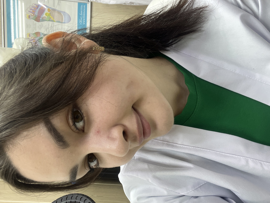 A young woman with dark hair and eyes, wearing a white coat over a green shirt, is angled slightly towards the camera. She has an earring in her left ear. In the background, a chart with colorful diagrams is visible. Her skin is light, and her expression is subtle, almost a slight smile. A portion of a circular object appears at the bottom left.