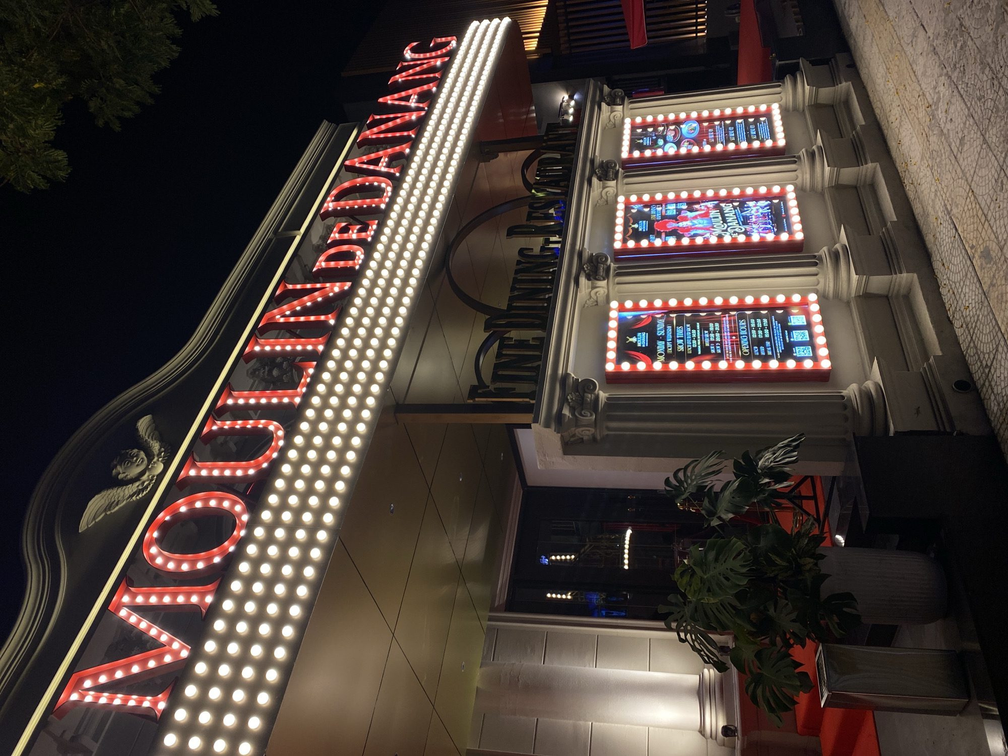 This is a night-time exterior shot of the Moulin Rouge de Danang, a fine dining restaurant located in Da Nang, Vietnam. The restaurant's facade is brightly lit with red neon letters spelling out "MOULIN de DANANG" and a marquee of incandescent bulbs. Below the main sign, smaller illuminated display boards showcase showtimes and menu items. The building features classical architectural elements, including columns and an ornate upper facade with a winged cherub motif. A red carpet leads to the entrance, and a large potted plant with broad green leaves is situated to the left of the entrance. The scene is set against a dark night sky, with the glow of the restaurant's lighting illuminating the paved sidewalk and surrounding street.