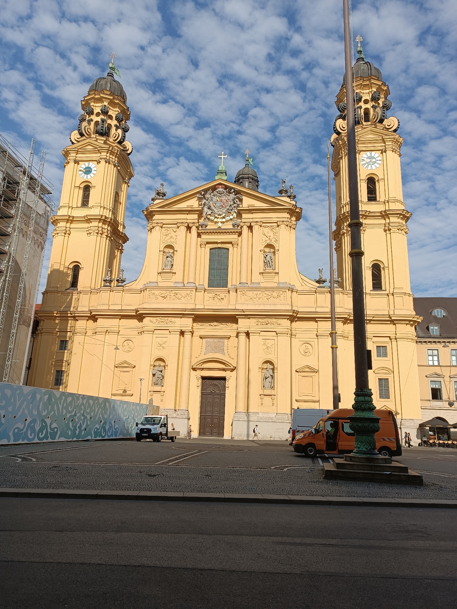 The image shows the Theatinerkirche in Munich, Germany. The church is a large, Baroque Roman Catholic church with two prominent bell towers. It is painted a distinctive yellow color. In front of the church, there is a paved square with cobblestones. Several vehicles are parked on the square, including a white van, an orange van, and a white truck. There are also a few people walking around the square. In the background, there are other buildings, including some with dark roofs. The sky is blue with scattered white clouds. Scaffolding is visible on the left side of the church, indicating ongoing construction or renovation. A large decorative fence with a repeating pattern is erected in front of a portion of the church.