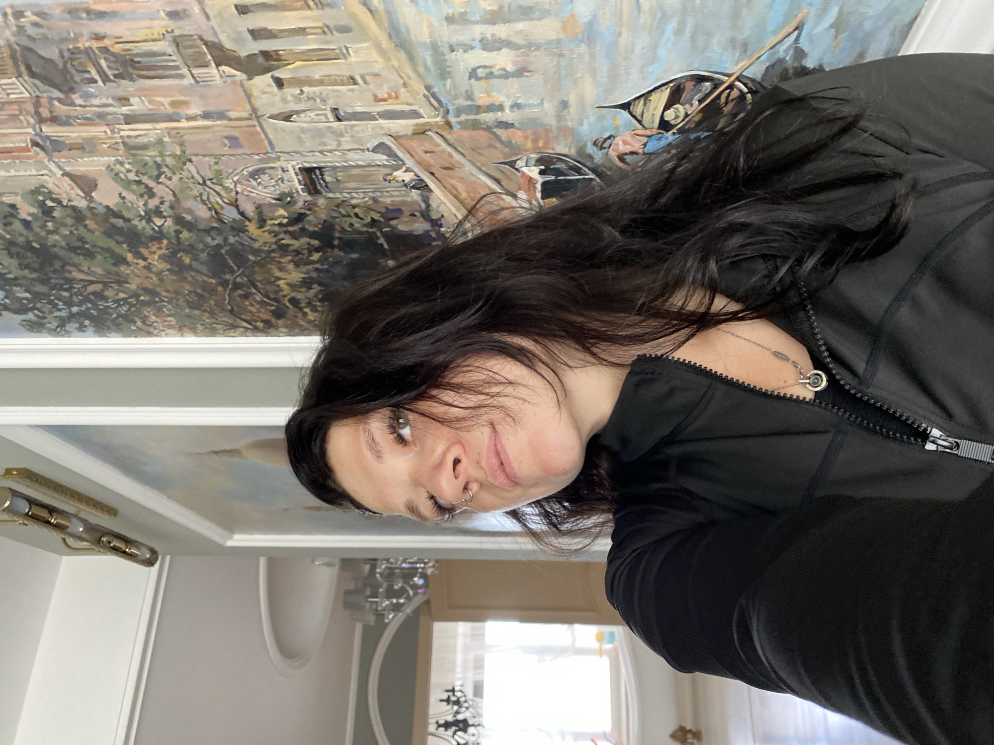 The image is a portrait of a young woman with long, dark hair, wearing a black jacket and a silver necklace. She has a nose ring and appears to be looking at the camera with a slight smile. The background is a painting of a Venetian canal scene, with gondolas and buildings. The lighting suggests it is daytime. The woman is positioned in the foreground, with the painting taking up the upper portion of the frame. The overall mood is casual and relaxed. There is no visible text in the image.