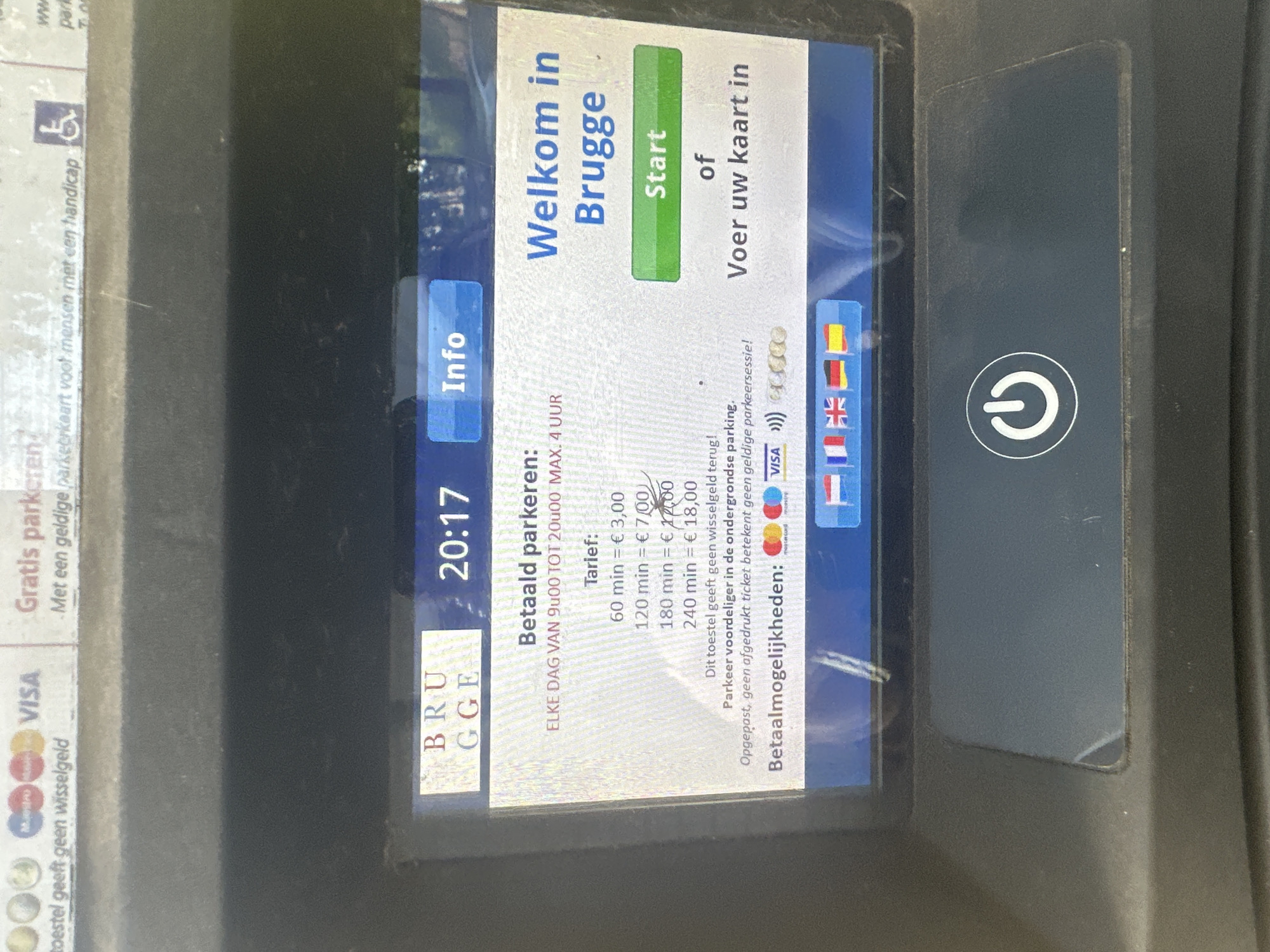 The image shows a close-up view of a parking meter screen in Bruges, Belgium. The screen displays "Welkom in Brugge" (Welcome to Bruges) and "Start" or "Voer uw kaart in" (Enter your card). The time displayed is 20:17. Below this, there is information about parking tariffs and payment methods, including Mastercard, Visa, and contactless payment symbols. The meter also shows flags representing different languages, including Dutch, French, English, German, and Spanish. The screen is slightly smudged, and some light reflections are visible. The surrounding machine is dark grey and appears to be made of plastic. A power button symbol is visible at the bottom of the screen. The overall impression is a functional parking payment terminal in a public area.