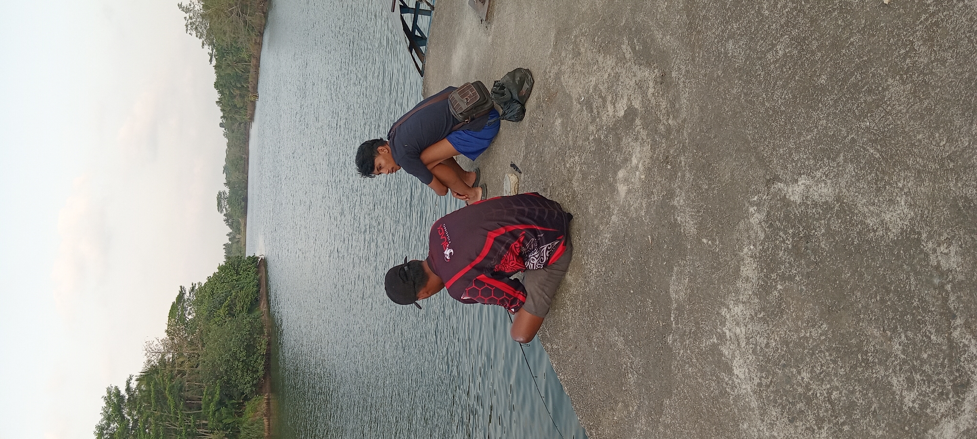 Two young men are sitting on a concrete embankment by a wide, calm river in Kalipucang, Indonesia. The man on the left, facing away from the camera, is wearing a black baseball cap and a maroon and black long-sleeved shirt with a red design. He is holding a fishing rod and appears to be fishing. The man on the right, facing left, is wearing a dark blue t-shirt, blue shorts, and sandals. He is looking out at the water and has a brown crossbody bag slung over his shoulder, and a dark green bag on the ground beside him. The river is bordered by lush green trees and vegetation on both sides, and the sky is overcast. The concrete embankment is rough and textured, with some debris scattered on it. The overall scene suggests a peaceful afternoon spent fishing by the river.