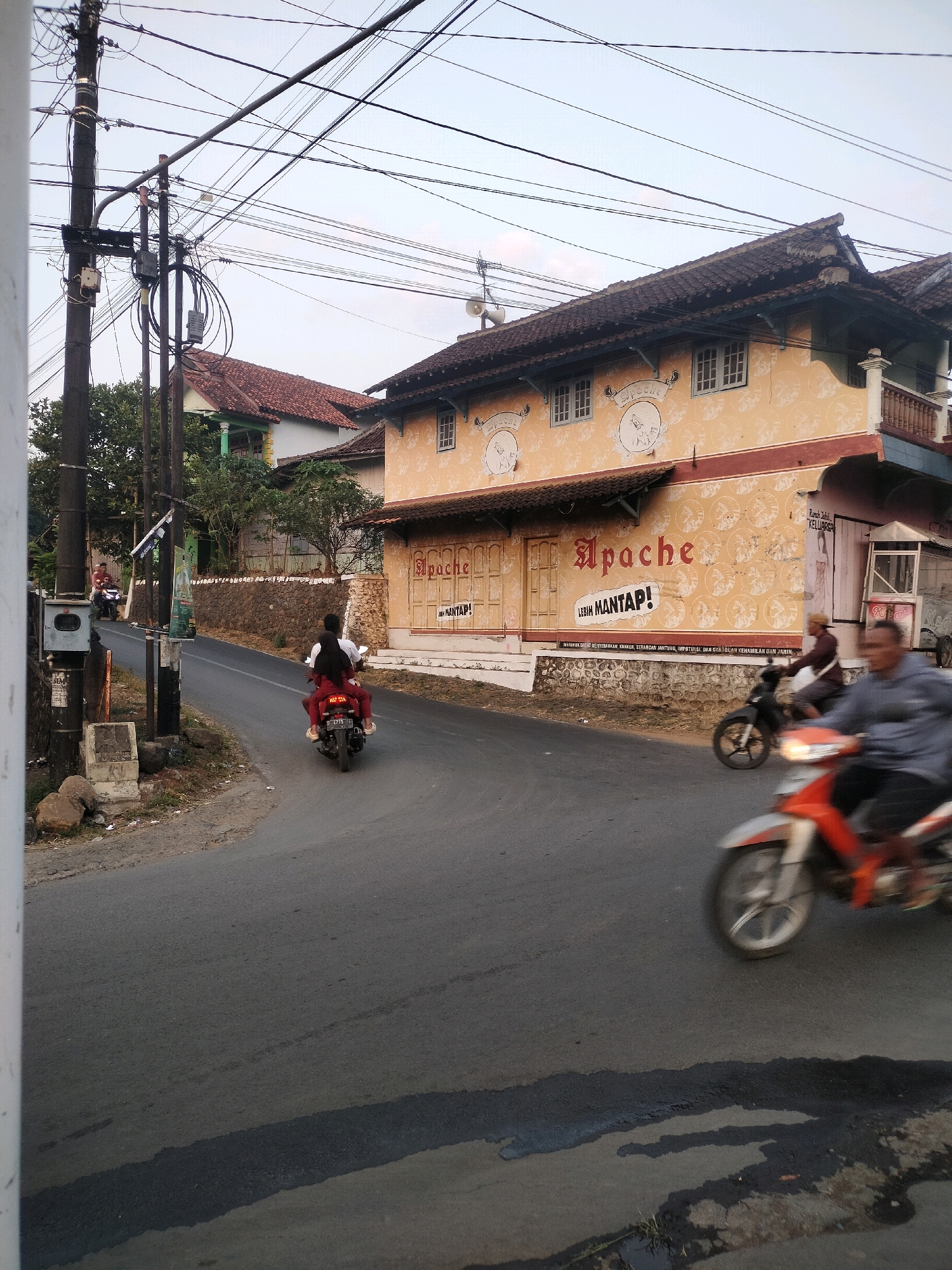 This is an eye-level, medium shot of a road in Boja, Indonesia, taken during the day. A paved road curves through a semi-rural area. On the right side of the road, a building with an orange facade and white decorative patterns stands. The building has a tiled roof, several windows, and a sign that reads "Apache" in a stylized font. There are multiple utility poles with tangled wires on the left side of the road. Several people are riding motorcycles. In the foreground, a person on an orange and white motorcycle is moving from right to left, blurred due to motion. Further down the road, two people are riding a black motorcycle from left to right. Another motorcycle is visible in the background on the left, with a rider. The environment appears somewhat aged, with visible wear on the road surface and some debris near the utility poles.