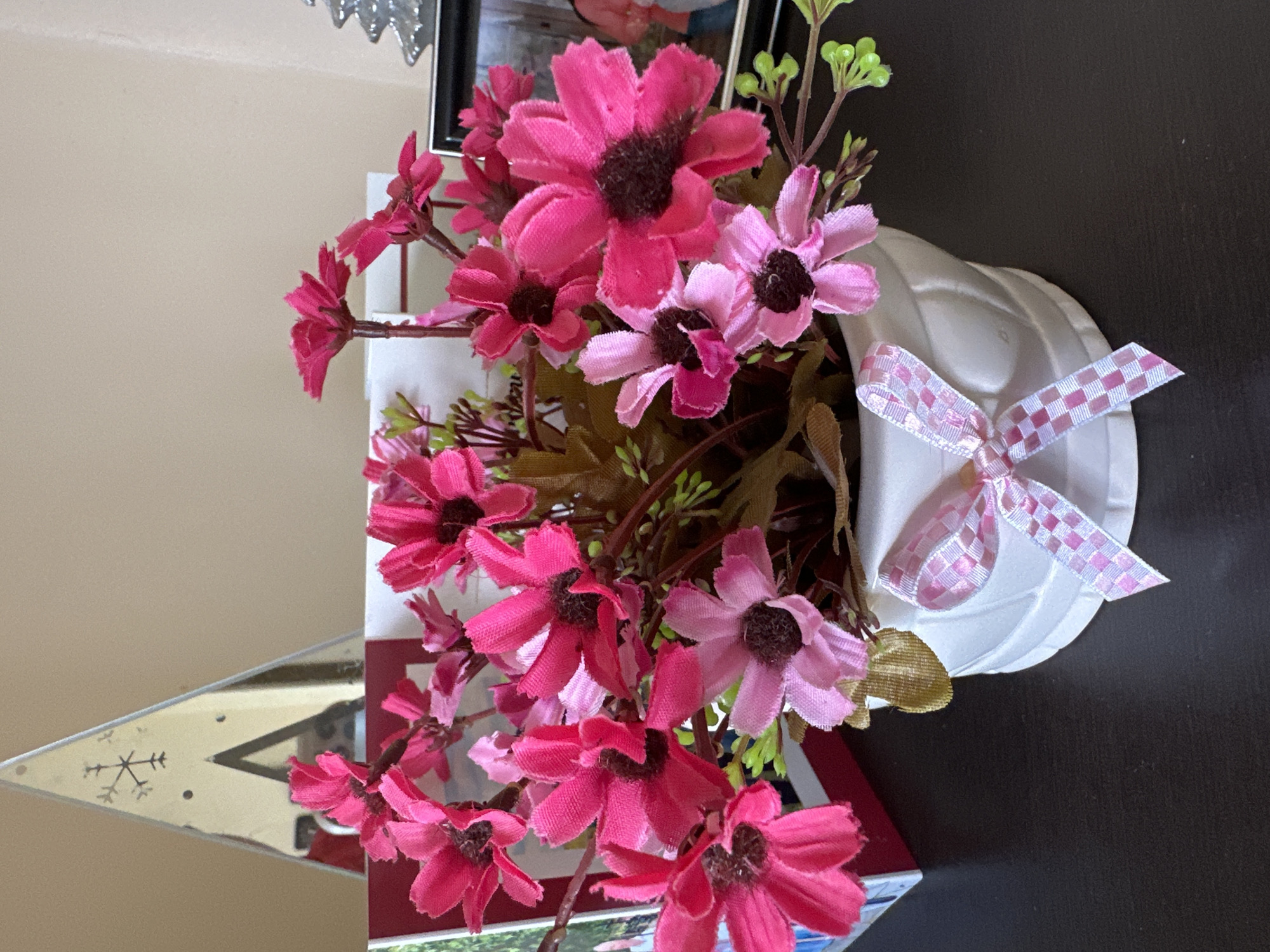 A floral arrangement in a white, heart-shaped vase is centrally located in the image. The flowers are primarily shades of pink and magenta, with dark brown centers. A pink and white checkered ribbon adorns the vase. The arrangement sits on a dark brown surface. In the background, blurred objects and a beige wall are visible. A decorative, triangular object with etched designs can be seen to the left. The image appears to be taken indoors. No information is available regarding the image's location within Rogozinovo, Bulgaria, or the presence of any people or specific actions.