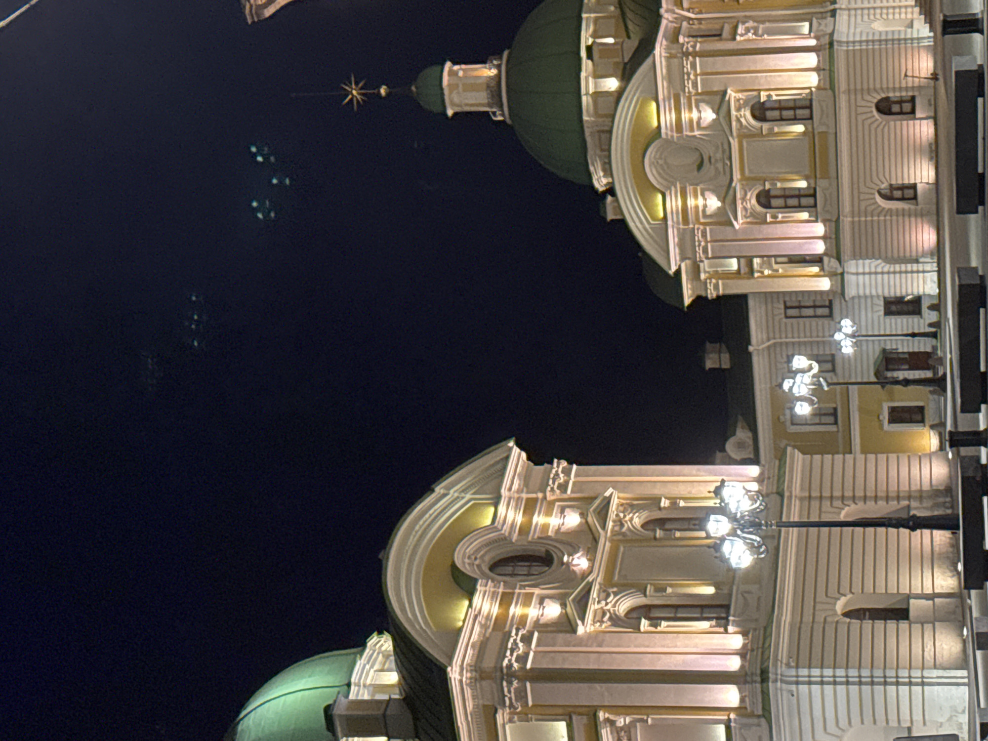 At night, an upward view of a building with green domes and illuminated beige facades dominates the scene. The architectural details feature ornate columns, arches, and windows, all lit up. Street lamps with multiple lights contribute to the brightness around the base of the building. The background is a dark, nighttime sky with some out-of-focus light spots, possibly from distant city lights. It is the Ascension Cathedral in Tver, Russia.