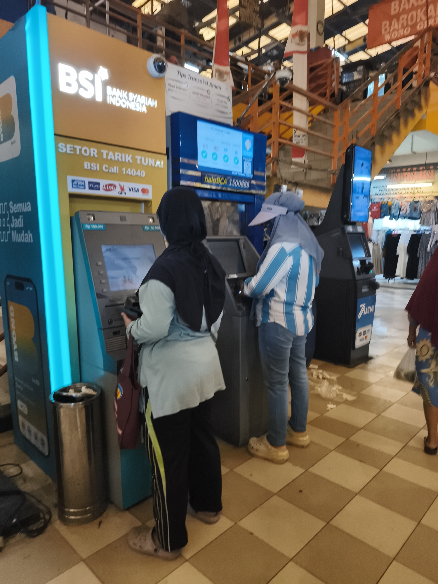 In North Jakarta, Indonesia, at what appears to be an indoor market or shopping center, two individuals are using ATMs. The ATM on the left, a blue and yellow BSI (Bank Syariah Indonesia) machine, has "SETOR TARIK TUNAI" (Cash Deposit Withdrawal) and "BSI Call 14040" displayed. It also shows logos for ATM Bersama, Prima, Link, GPN, and Visa. The ATM on the right is black with a prominent "ATM" logo and Visa branding. Both individuals are dressed modestly, with one person wearing a headscarf and the other a head covering and a blue and white striped shirt. The environment features tiled flooring, overhead structures, and signage, including a red sign with white lettering reading "BAROKAH WONO GIRI". Clothing racks with garments are visible in the background, suggesting a commercial setting. A metal trash bin is situated next to the BSI ATM.