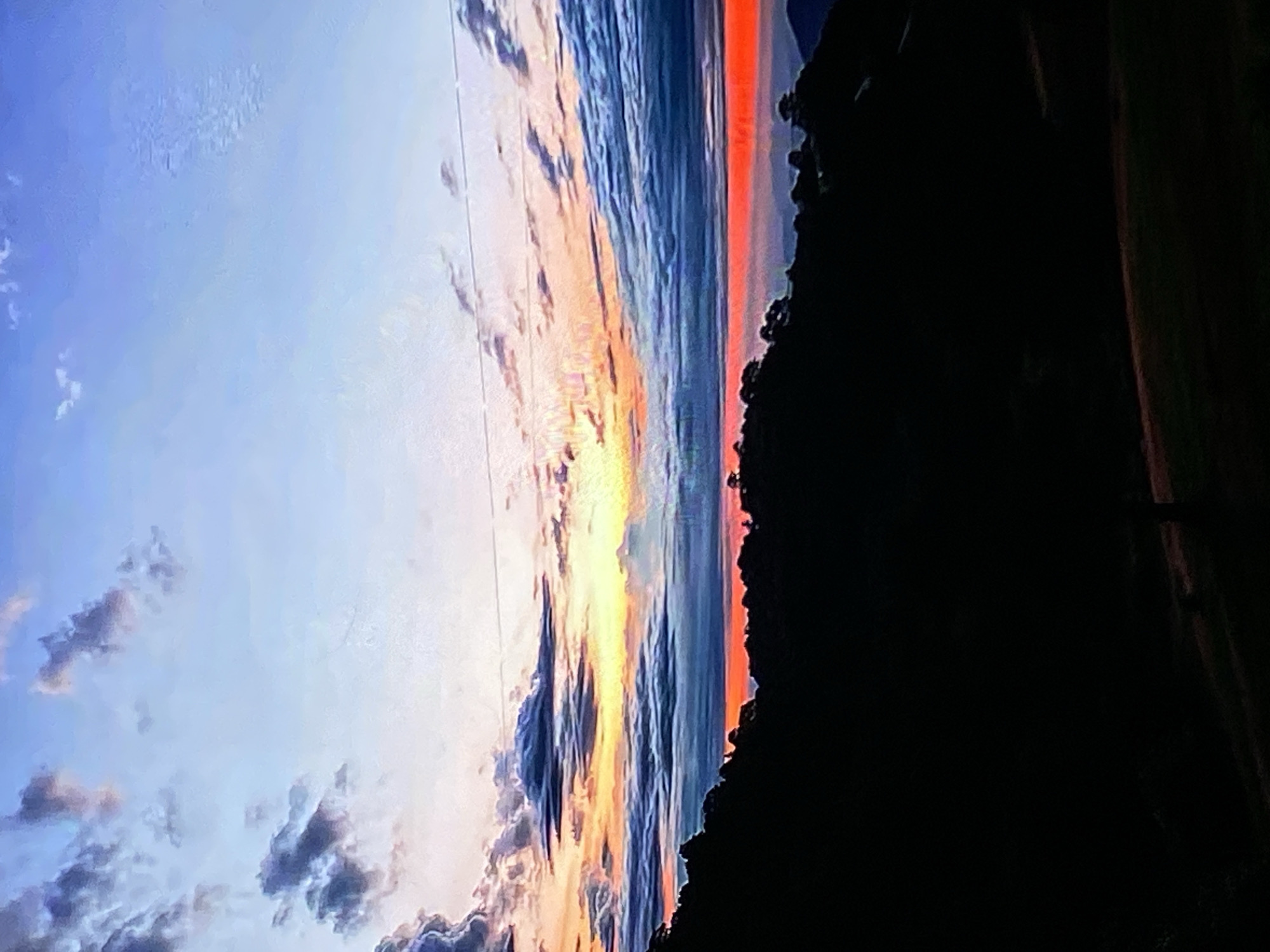 The image displays a panoramic view of a twilight sky over a mountainous landscape in Tinaquillo, Venezuela. The sky is a vibrant gradient of blues, oranges, and yellows, with scattered clouds. The foreground is dominated by the dark silhouette of a mountain range, sparsely dotted with trees. A thin, bright orange band runs horizontally across the horizon, marking the transition between the sky and the land. Two thin, horizontal lines, possibly power lines, stretch across the upper portion of the sky. The overall impression is of a serene and natural landscape at dusk.