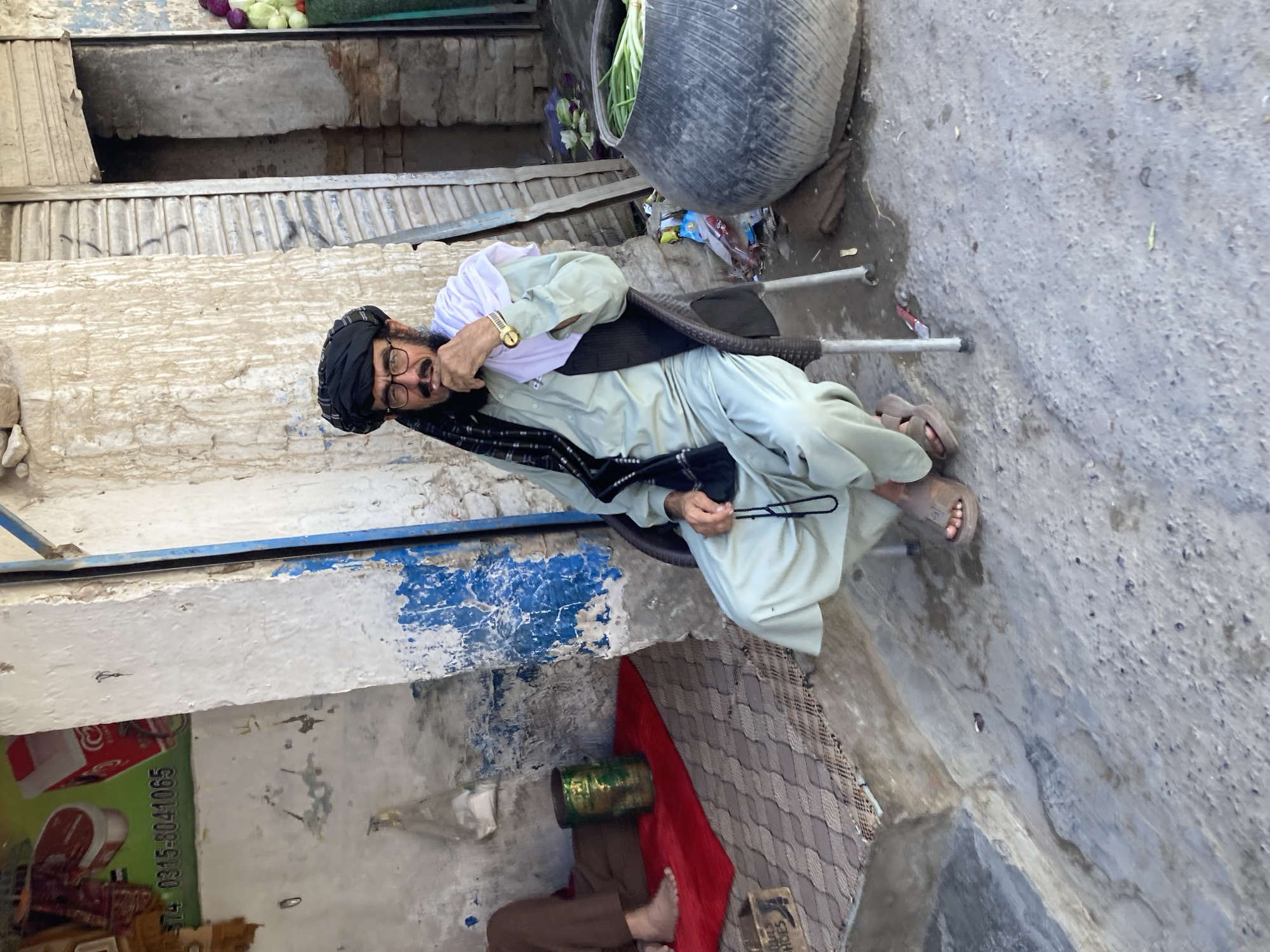An elderly man sits on a plastic chair in an alley in Quetta City Tehsil, Pakistan. He wears a black turban, glasses, a white shawl, and a light green shalwar kameez with a black vest. He holds a black prayer bead string. His sandals are visible on his feet. He is looking towards the camera with his mouth open. The alleyway is narrow, with buildings lining both sides. One building has a food advertisement on it, with the phone number "0315-8041065" visible. A bed with a red blanket is underneath him, with a green drum next to the bed. Debris and garbage are strewn on the ground. A large tire sits at the end of the alleyway.