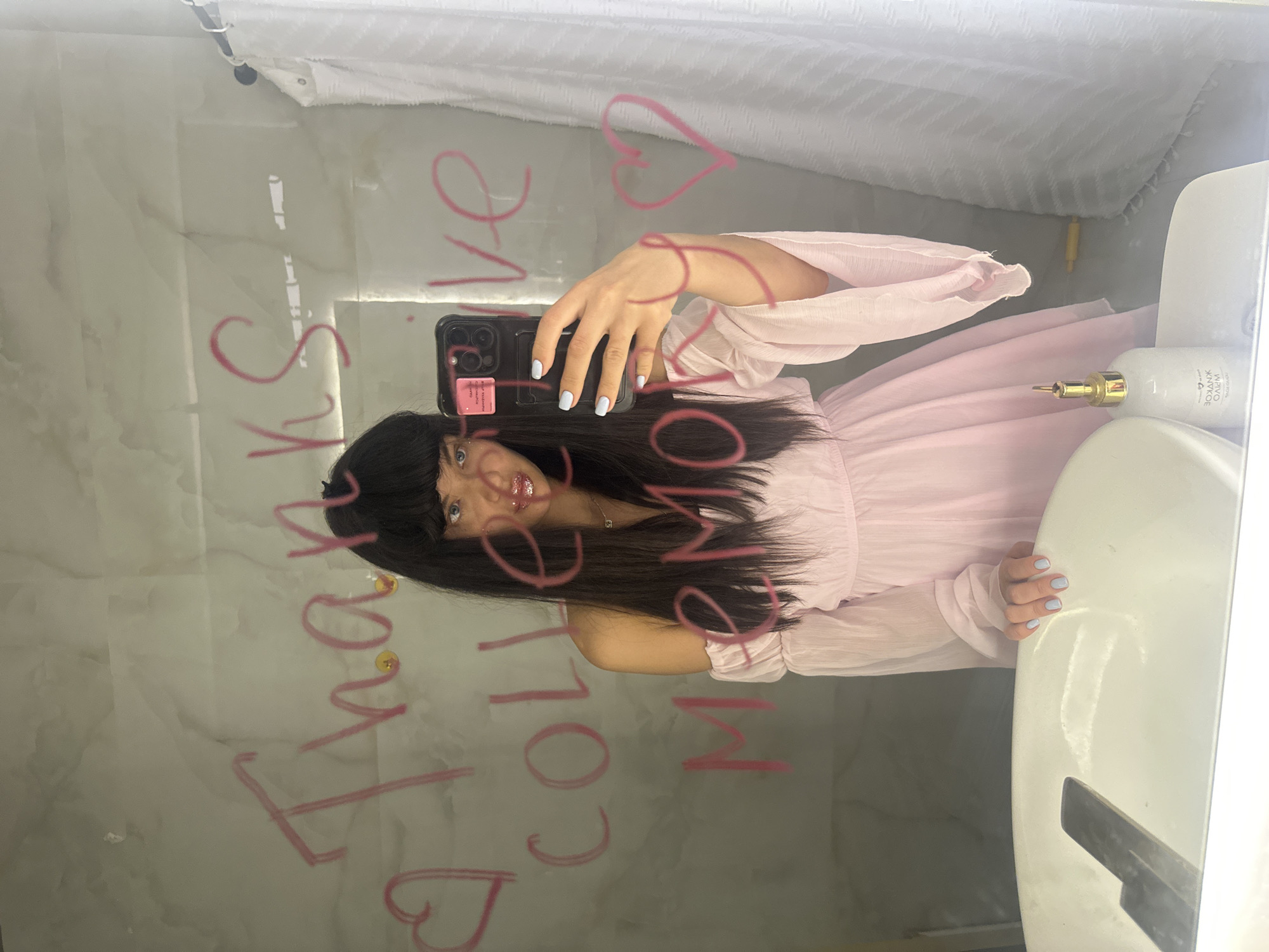 The image displays a mirror selfie of a young woman in what appears to be a bathroom, captured from a rotated perspective. The woman has long, dark hair with bangs and light blue eyes. She is wearing a light pink dress with long, sheer sleeves and a gathered bodice. Her fingernails are painted a soft light blue. She holds a black smartphone with multiple camera lenses in her right hand, partially obscuring her face as she takes the photo. A pink sticker or attachment is visible on the back of her phone case. She has a slight smile and her lips are adorned with glittery gloss.
Prominently written in pink on the mirror's surface is the message: "Thanks for follow me ❤️". This message is scrawled in a casual, handwritten style.
Reflected in the mirror behind the woman is a light-colored wall with a subtle marble pattern, and above it, a white, textured fabric, likely a shower curtain or a similar textile wall covering. To the right of the frame, a white sink basin is visible. On the sink's edge, there are two bottles: one with a golden pump dispenser and another taller white bottle, which has partially legible text on its side.
The activity depicted is a casual mirror selfie, with a personalized message added to the mirror. The setting is an indoor space, strongly suggested to be a bathroom due to the presence of a sink and what appears to be a shower curtain. The overall tone is light and appreciative, as conveyed by the written message.