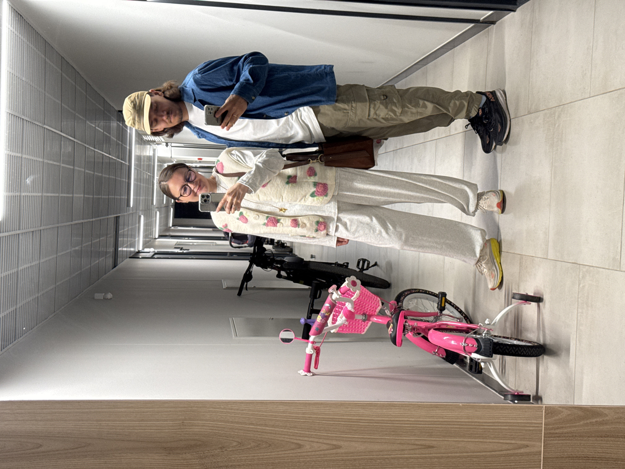 The image shows two individuals, a man and a woman, standing in a brightly lit indoor hallway. Both are looking at their smartphones.
The man, positioned on the left, wears a light brown baseball cap, a blue denim-like jacket over a white t-shirt, and olive green cargo pants. He has long, dark hair and a ring on his right ring finger. His dark running shoes feature a white sole with the brand name "HOKA" visible on the side.
The woman, to the man's right, wears glasses, a light-colored long-sleeved top, and a cream-colored vest with embroidered or appliquéd pink roses. She has light-colored pants and light-colored athletic shoes with yellow and orange accents. A brown leather shoulder bag hangs from her left shoulder.
Parked on the tiled floor to their right are two bicycles. The foreground features a vibrant pink child's bicycle equipped with training wheels and a white front basket. Behind it, a darker, possibly black, adult bicycle is partially visible. The hallway has a light-colored tiled floor and a wall with a grid-like texture or reflective panels on the left. The bottom of the frame shows a wooden floor or wall panel. The scene appears to be taking place indoors, with artificial lighting, suggesting it could be day or night.