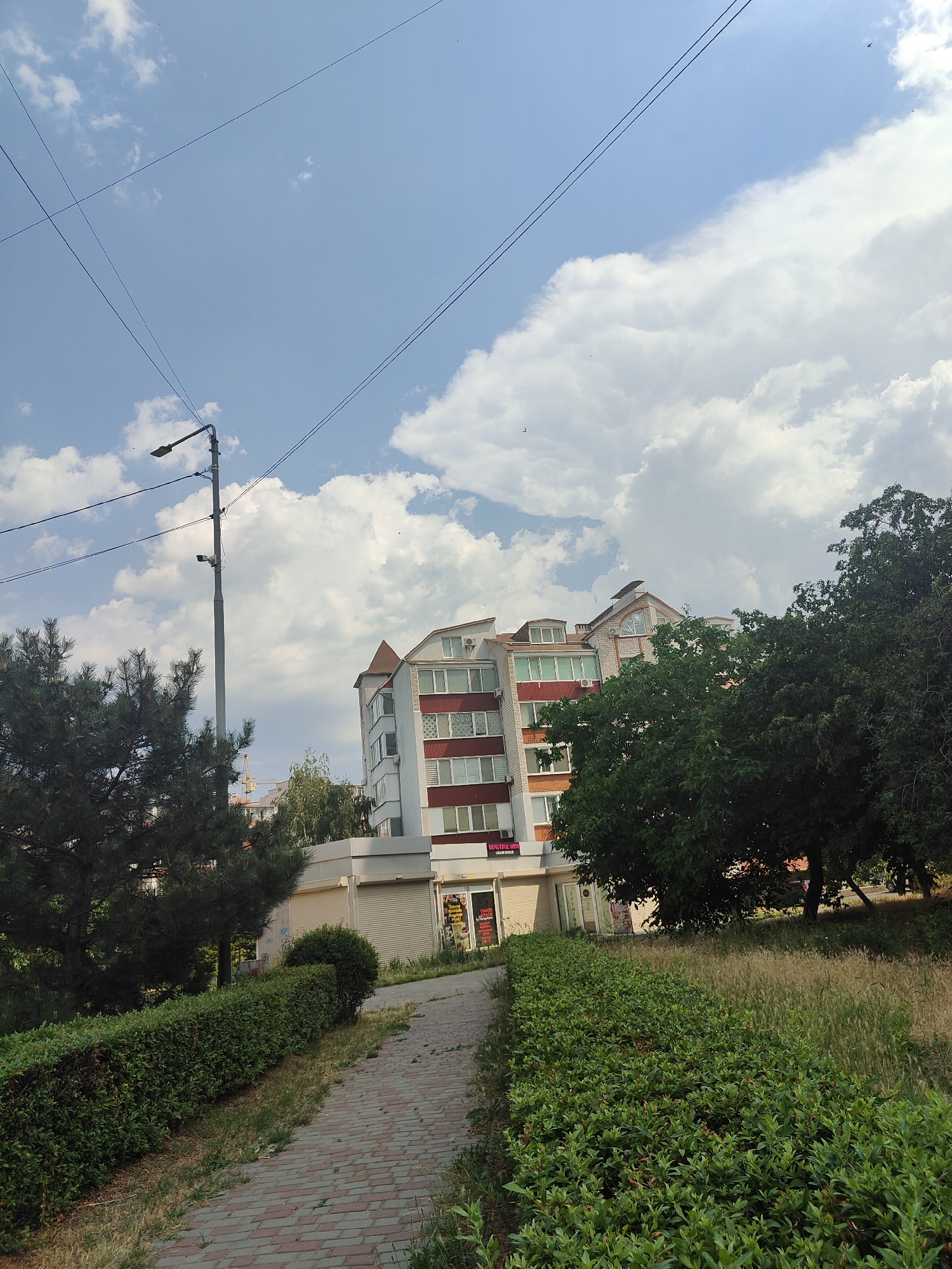 The image depicts a view of a modern building with a red and white facade, surrounded by greenery under a bright, partly cloudy sky. A paved pathway winds through manicured bushes in the foreground, leading towards the building. To the left of the pathway, a tall pine tree stands, and a lamppost with a security camera is visible. A larger, leafy green tree is situated to the right of the building. In the distance, construction cranes suggest ongoing development. The sky is a vibrant blue with scattered white cumulus clouds, indicating fair weather. The lighting suggests it is daytime, likely mid-morning or afternoon. Visible text on the building facade includes "BEAUTIFUL" and what appears to be "SALON". The scene appears to be a residential or commercial area in Chornomorsk, Ukraine. No people are visible.