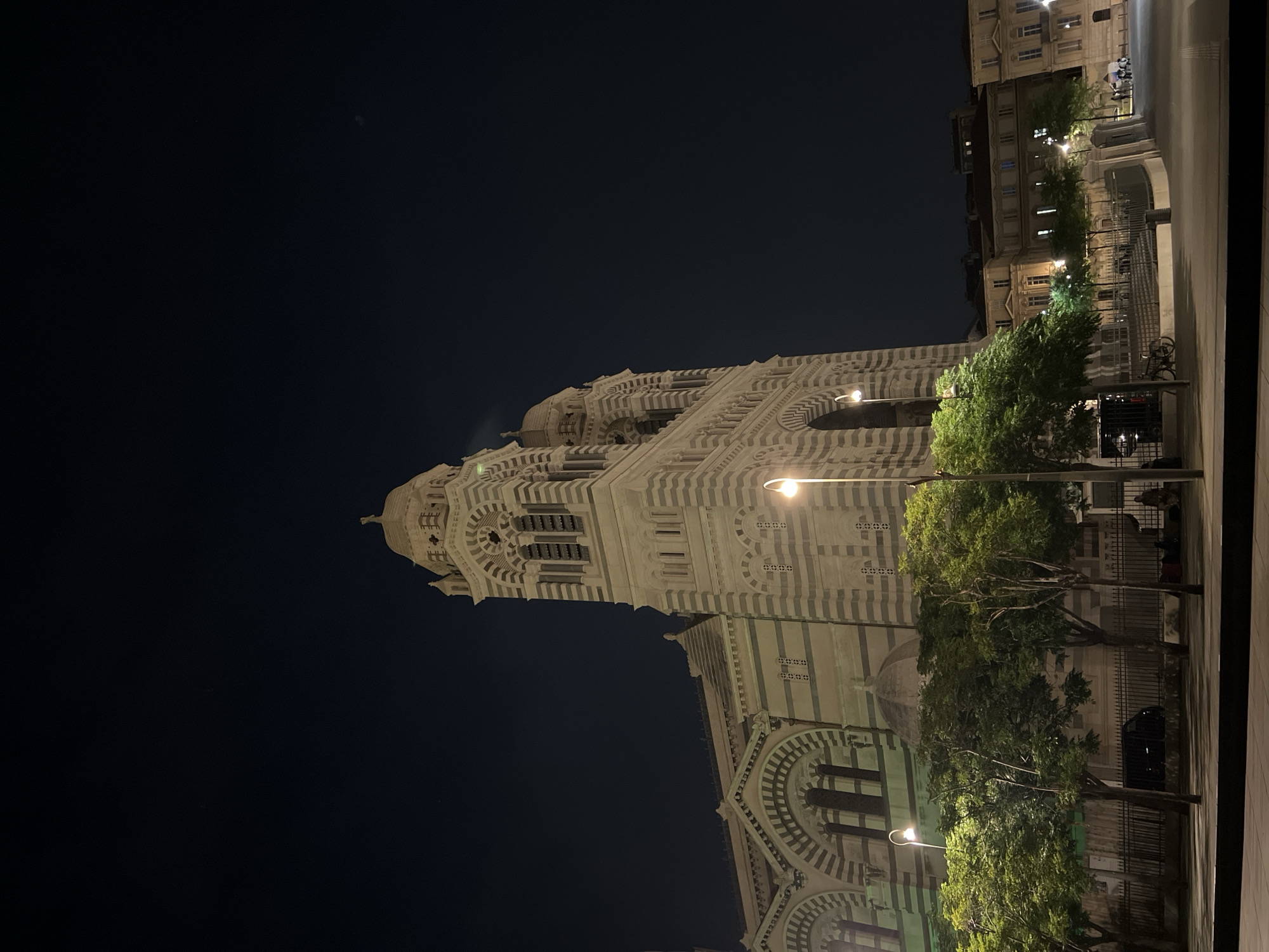The image depicts a nighttime view of the Cathédrale La Major in Marseille, France. The cathedral, with its distinctive striped facade and twin towers, is illuminated by streetlights. In front of the cathedral, there are several trees casting shadows on the paved ground. Some people are visible sitting or standing in the background, near the entrance to the cathedral, and a black car is parked behind a fence. The sky is dark, indicating it is nighttime. The scene is calm and serene.