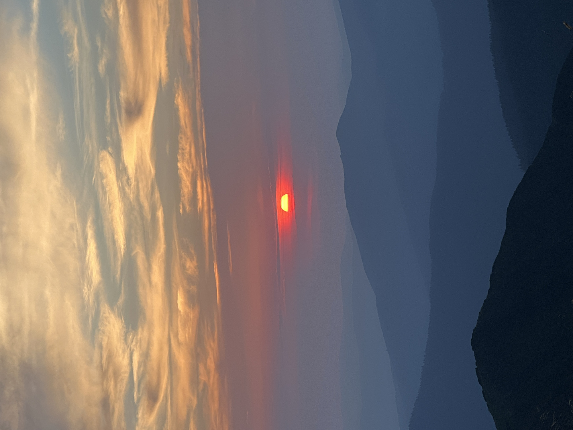 A serene mountain landscape is captured at sunset. The sky is painted with a gradient of colors, from warm oranges and yellows at the top to soft pinks and purples near the horizon. Wispy clouds stretch across the sky, illuminated by the fading sunlight. A large, vibrant red sun is partially obscured by haze, positioned in the middle of the frame, casting a warm glow. Below the sky, several layers of mountains recede into the distance, depicted in progressively darker shades of blue and gray, creating a sense of depth. The foreground mountain is the darkest and most detailed, with a jagged silhouette. The image does not contain any people or recognizable landmarks that would identify it as being in the city of Luhy, Ukraine.