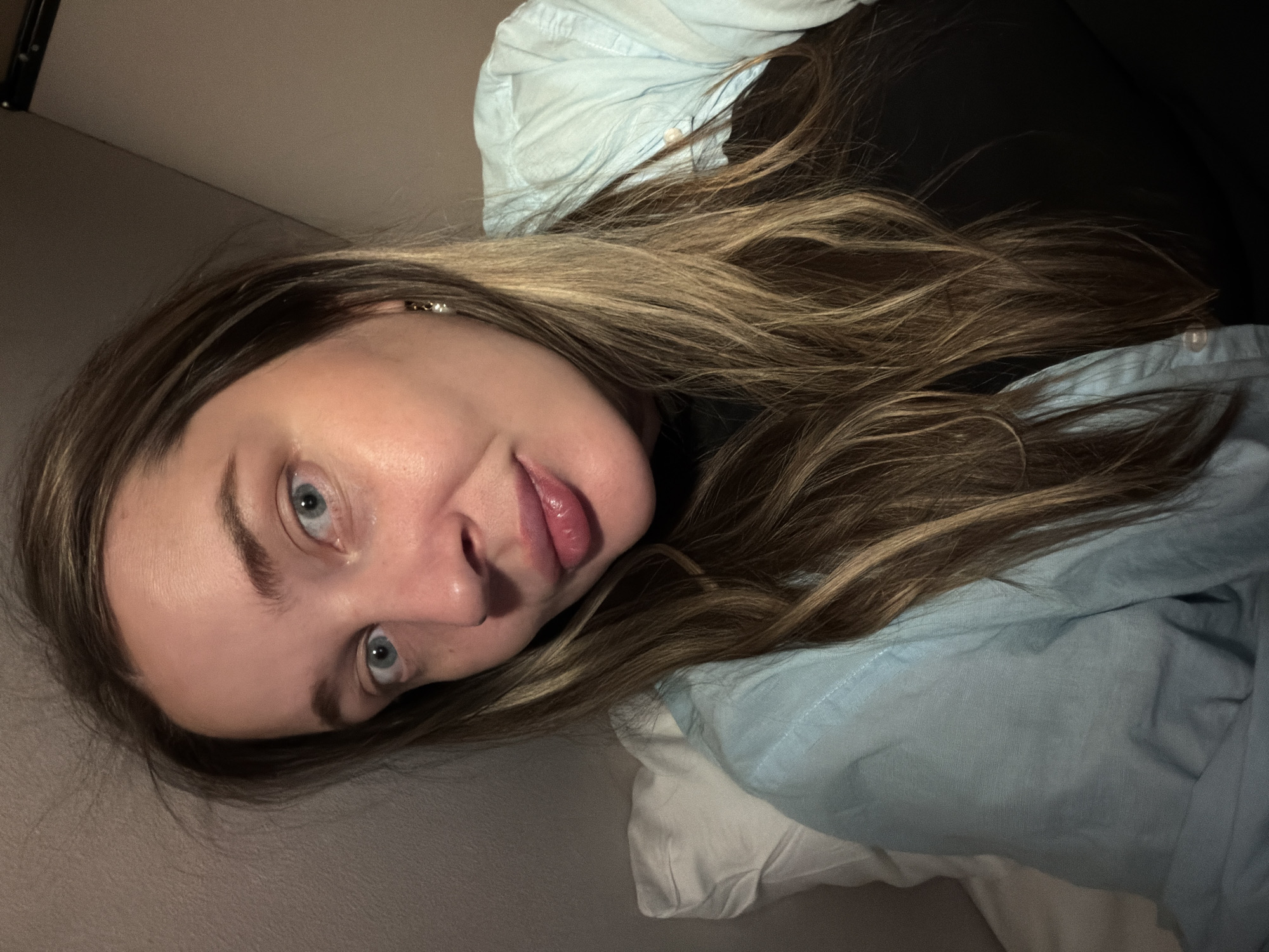 A woman with blonde and brown hair is lying on a white and blue pillow on what appears to be a bed. She has blue eyes and her mouth is slightly open, sticking out her tongue. The background is a dark, solid color, possibly a wall. There is no evidence to suggest the location is Moscow, Russia.