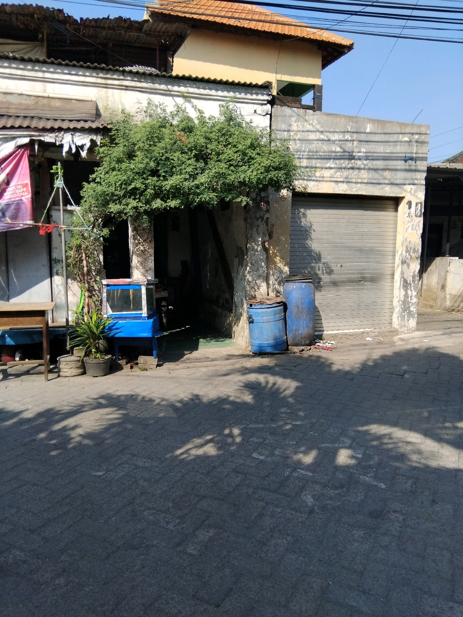The image captures a scene in Kepuh Kiriman, Indonesia, under a clear blue sky with shadows from trees cast across the textured pavement. In the foreground, the pavement is made of interlocking grey bricks. To the left, a simple wooden table with one leg is positioned. Further back, a blue and silver display case with glass panels stands on the pavement, accompanied by two potted plants and a slender tree.
Behind the display case, an opening leads into a dimly lit interior. A large, mature tree with dense green foliage grows prominently in front of this opening, obscuring some of the building's facade. To the right of the opening, two large blue plastic barrels are situated on the pavement; one is taller and the other is shorter and wider. The building itself features a textured, weathered concrete facade. A grey, corrugated metal roll-up door occupies a significant portion of the right side of the building. Above the roll-up door, the number "10" is visible in faded yellow paint. The building has a light-colored, textured upper wall and a reddish-tiled roof. Wires are strung across the upper portion of the scene. A pink banner, partially visible on the far left, hangs from the building.