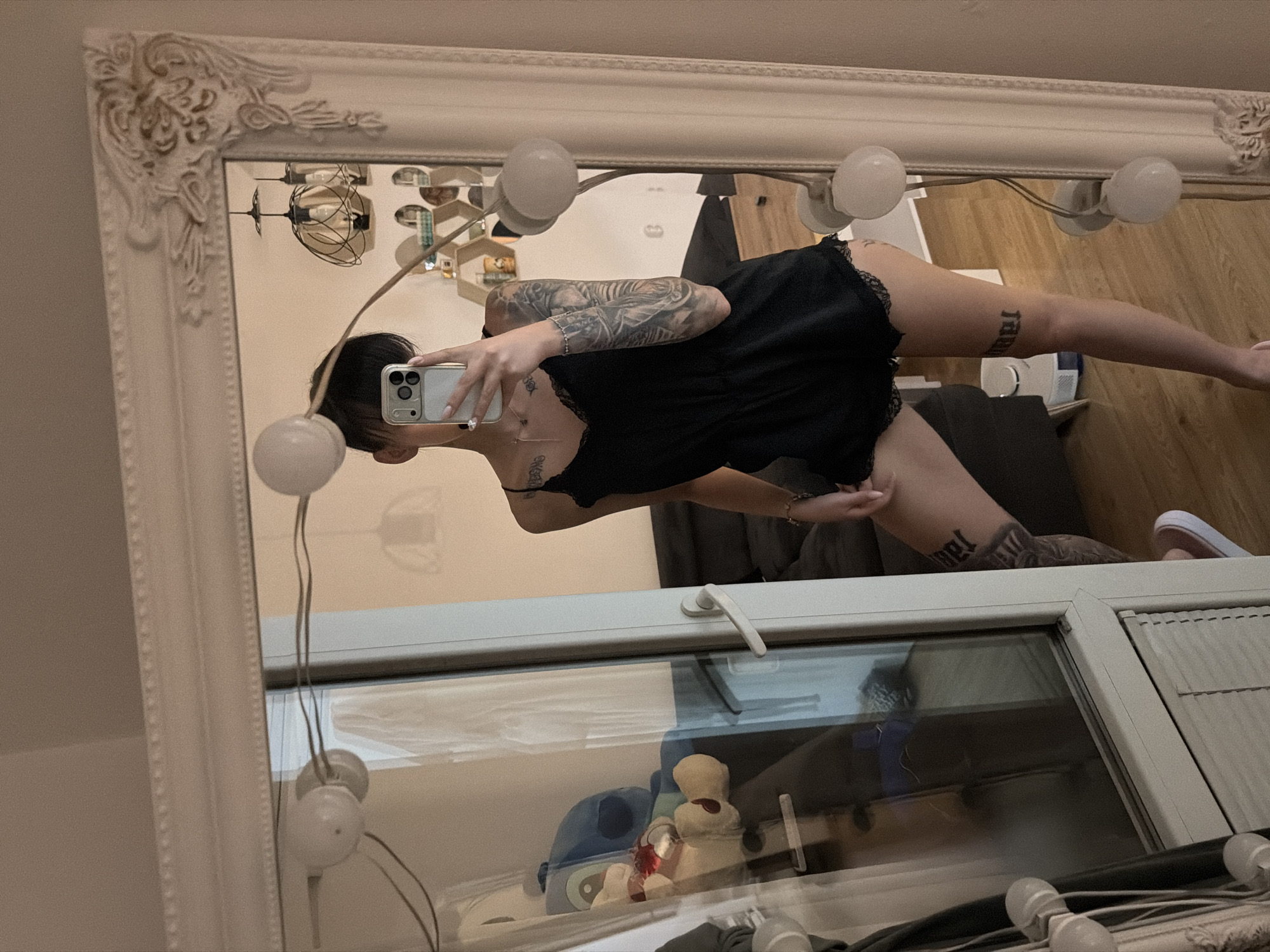 The image shows a young woman with dark hair taking a selfie in a full-length mirror. She is wearing a black lace-trimmed romper and has tattoos on her arms and legs. The mirror is ornate and framed in white. In the reflection, we can see a room with wooden floors, a sofa, and a stuffed toy on a shelf. The scene appears to be in a domestic setting, likely an apartment. There is no specific information to indicate the location is in Plovdiv, Bulgaria.