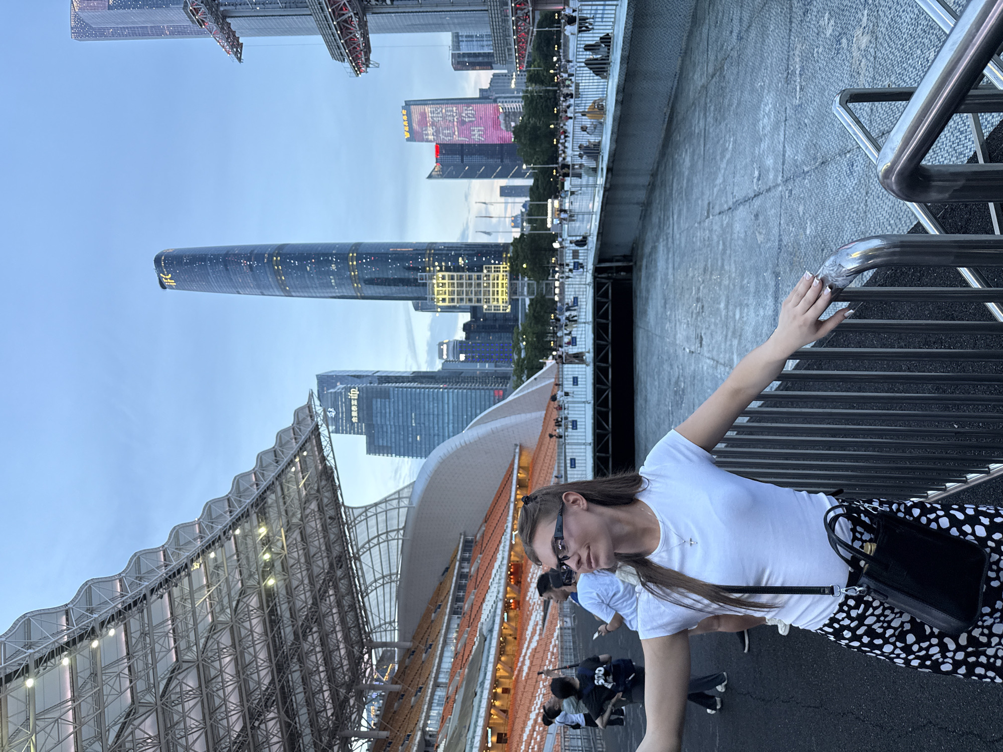 The image captures a woman in the foreground at dusk in the Tianhe District, China, likely near the Guangzhou International Finance Center. She is wearing a white t-shirt, a black and white spotted skirt, and glasses, and has a black crossbody bag. Her left arm is extended as if taking a selfie. Behind her, the distinctive wavy roof structure of a stadium, possibly the Guangzhou Stadium, is visible. In the background, the illuminated skyscrapers of the city, including the prominent IFC tower, stand against a twilight sky. Other people are scattered in the middle ground, some walking and some standing near the stadium seating. The environment appears to be an outdoor public space with metal railings and paved ground.