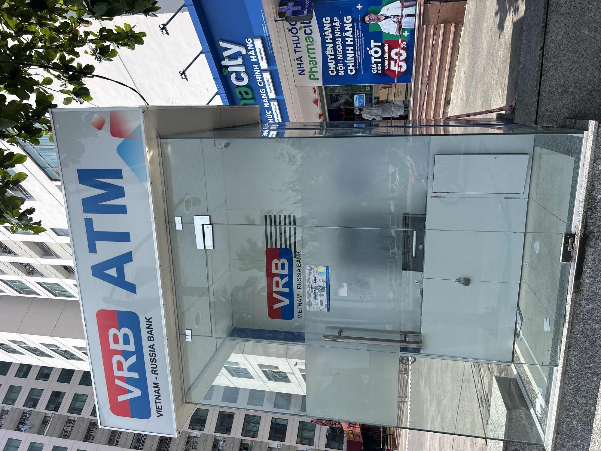The image shows a standalone VRB ATM (Vietnam-Russia Bank) booth in Bắc Nha Trang, Vietnam. The booth is made of glass and metal, reflecting the surrounding urban landscape. Above the booth is a sign with the VRB logo and the word "ATM". Adjacent to the ATM booth is a blue sign for a "Pharmacity" pharmacy. The pharmacy's promotional sign indicates a discount of up to 50% on certain items. There is a low wall/barrier near the steps leading into the pharmacy. The reflection in the glass shows buildings and other objects in the vicinity. The ATM booth appears to be empty.
