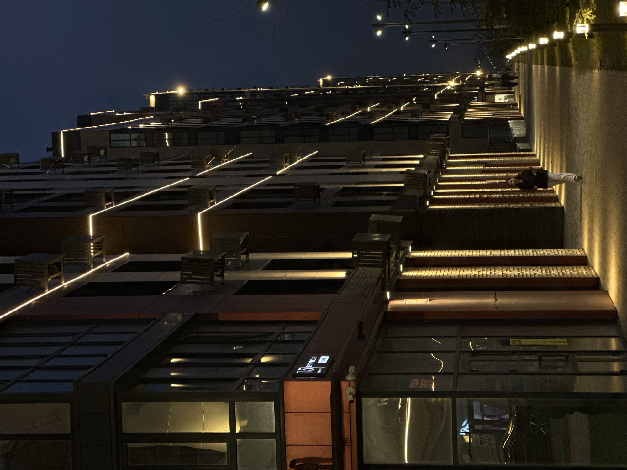 This image depicts an urban nighttime scene in Rostov-on-Don, Russia. The primary subject is a row of modern, multi-story buildings stretching into the distance along the left side of the frame. These buildings feature dark facades, large glass windows, and prominent architectural lighting consisting of horizontal LED strips along edges and vertical wall-washer lights illuminating ground-level brick columns. A visible sign on the lower-left building displays the number "15" and Cyrillic text, including "РОСТОВСКАЯ" (Rostovskaya), confirming the location.
A wide, paved pedestrian pathway runs parallel to the buildings. It is illuminated by the building's ground-level lighting and a series of short, square-shaped path lights positioned along its right edge. Further right, taller streetlights with multiple fixtures provide additional illumination.
One individual, appearing female, walks towards the foreground on the pathway, dressed in a dark jacket and light-colored pants. Her head is slightly turned left. Two or more indistinct figures are visible walking away from the foreground further down the path. To the right of the path is a landscaped strip containing small trees and bushes. The sky is dark blue-black, indicating late evening or night.