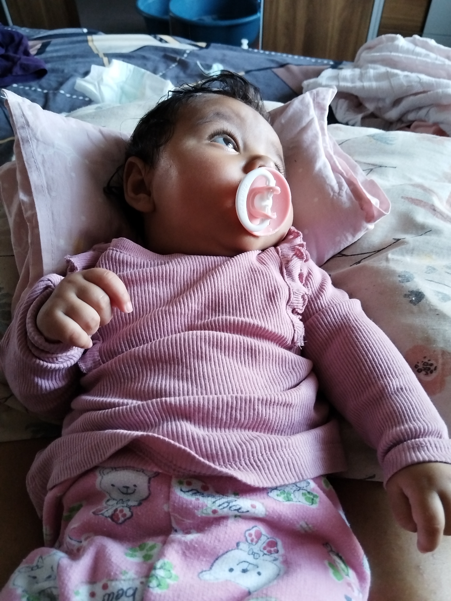 A baby wearing a pink, ribbed long-sleeved shirt and pink pajamas with teddy bear designs is lying on a bed with patterned bedding. The baby has a pink pacifier in their mouth and is looking upwards to the right. A pink pillow is under the baby's head. A blue bucket is visible in the background. The location is unknown.