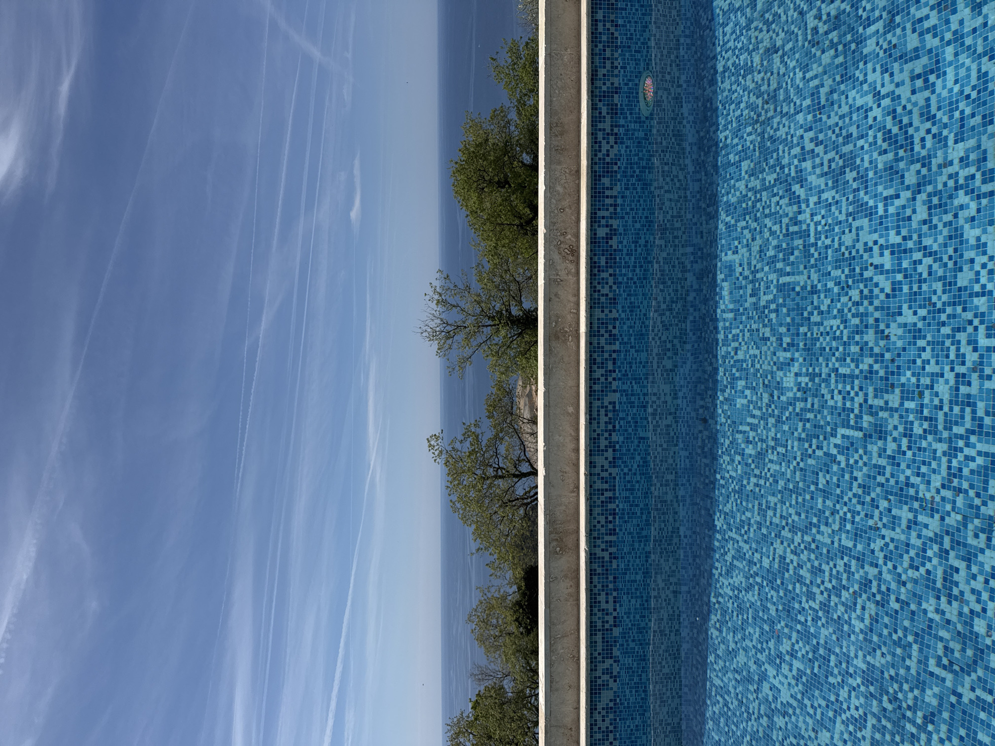 Captured from an elevated perspective in Blizikuće, Montenegro, the photograph presents a scene divided into three distinct vertical segments. The left segment showcases a clear, light-blue sky marked with contrails, suggesting recent air traffic. Adjacent to the sky, the central segment features a seascape merging with the horizon, and a line of trees along the coast. The right segment presents a swimming pool, its interior lined with blue mosaic tiles. The concrete pool edges run parallel to the seascape. There are no people visible in the shot.