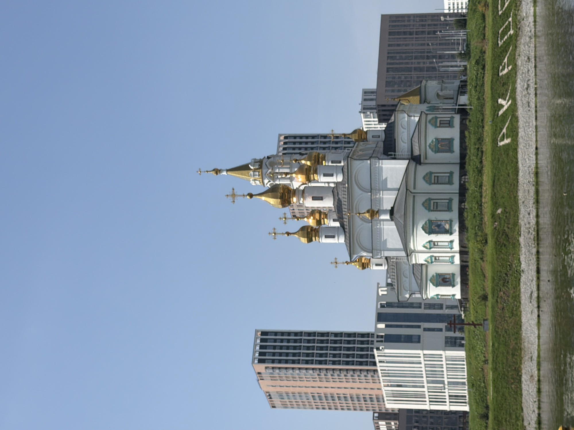 The image depicts a modern cityscape in Yekaterinburg, Russia, with a prominent Orthodox church situated in the foreground. The church, characterized by its white facade and multiple golden domes topped with crosses, stands on a grassy embankment overlooking a body of water. Behind the church, several contemporary high-rise buildings with glass and concrete exteriors dominate the skyline, representing the urban development of the city. The sky is clear and blue, suggesting a sunny day. On the grassy slope, the word "AKADEM" is spelled out in white letters, possibly indicating a district or institution. The overall scene captures a blend of historical religious architecture and modern urban infrastructure.