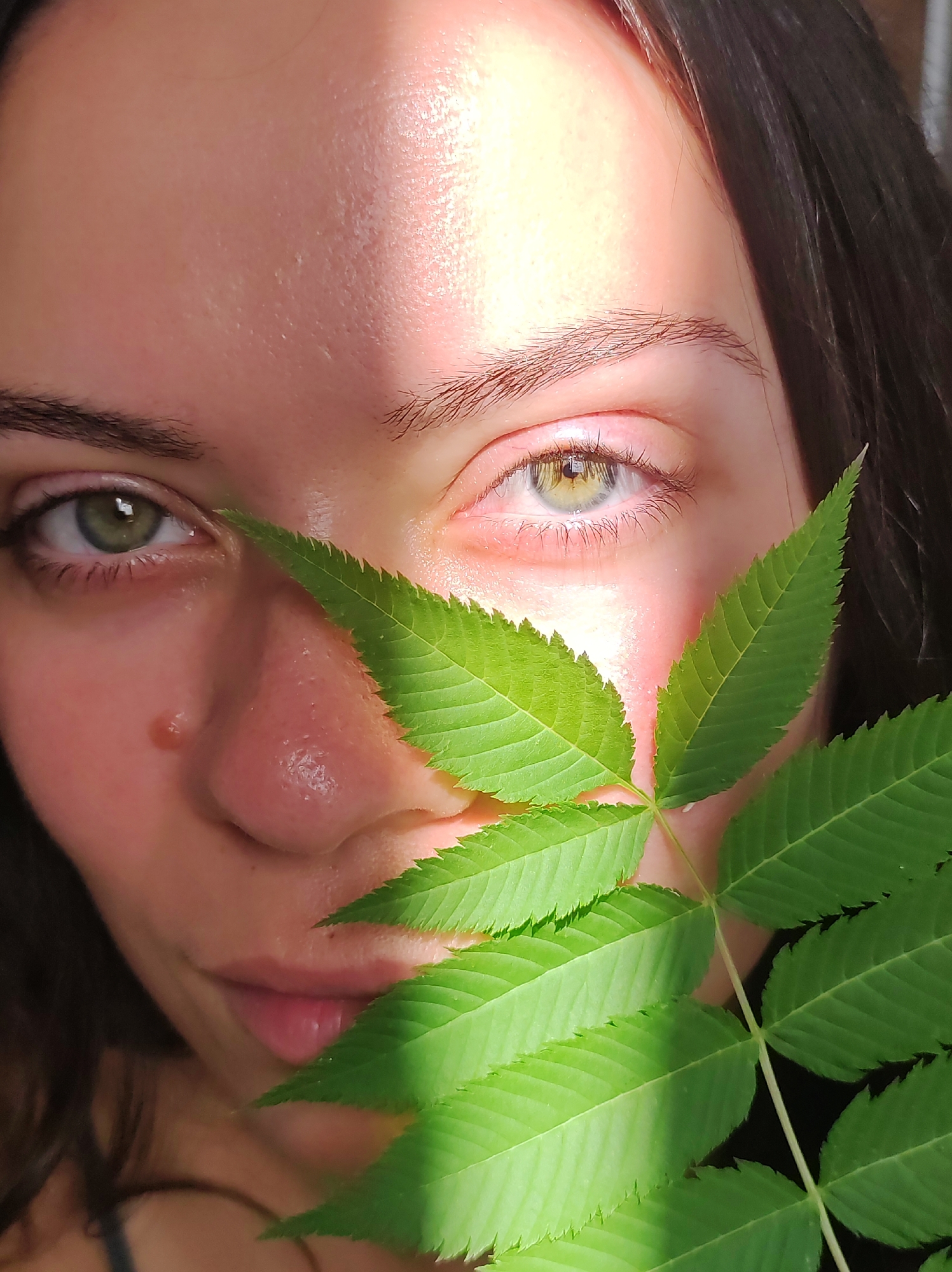 The image is a close-up portrait of a young woman's face, partially obscured by a branch with green leaves. The woman has fair skin, green eyes, and dark hair. A strong light source casts a shadow across her forehead and down her face. A dewdrop is visible on her cheekbone. The leaves are vibrant green with serrated edges. The background is dark and out of focus. There is no indication of the location being in Chaikovsky, Russia.