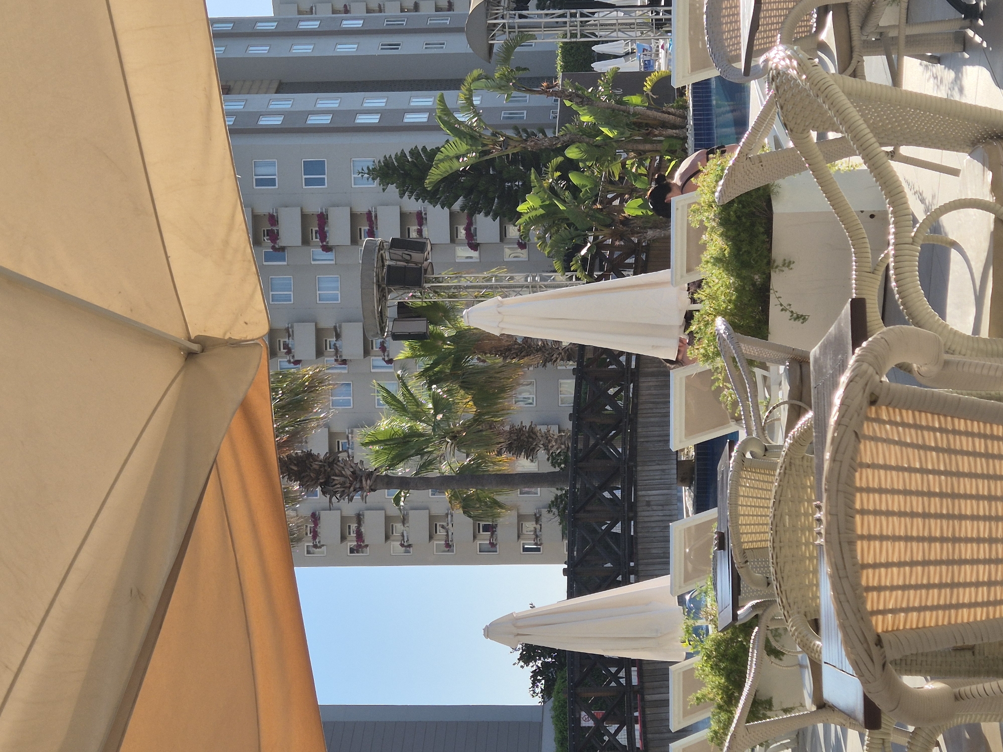 A resort swimming pool area is depicted in this medium shot, with a large umbrella in the foreground providing shade. White woven chairs and tables are arranged on the tiled deck. In the middle ground, a wooden fence borders the pool, beyond which are lounge chairs and lush green plants. A person in a black swimsuit is partially visible by the pool, facing away from the camera. In the background, a multi-story building with numerous windows and balconies, adorned with red flowers, stands tall under a clear blue sky. Palm trees and other tropical foliage are scattered throughout the scene, suggesting a warm climate. The overall atmosphere is one of leisure and relaxation.
