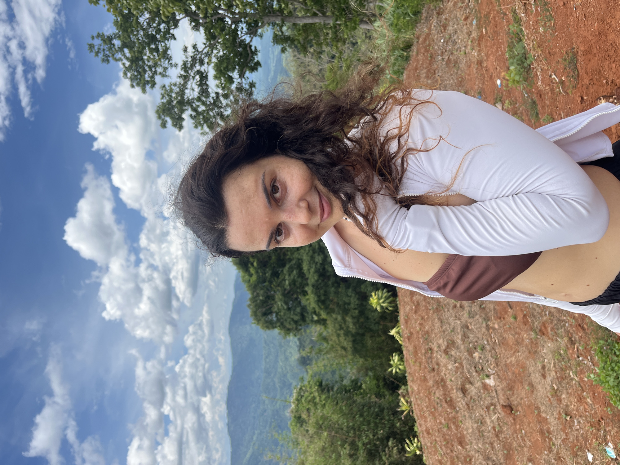The image shows a woman posing for a photo outdoors. She has long, curly brown hair and is wearing a brown bikini top and a white zippered jacket. She is looking at the camera with a slight smile. The background is a lush, green landscape with mountains and a cloudy blue sky. The ground in the foreground is reddish-brown soil. The location appears to be a scenic viewpoint.