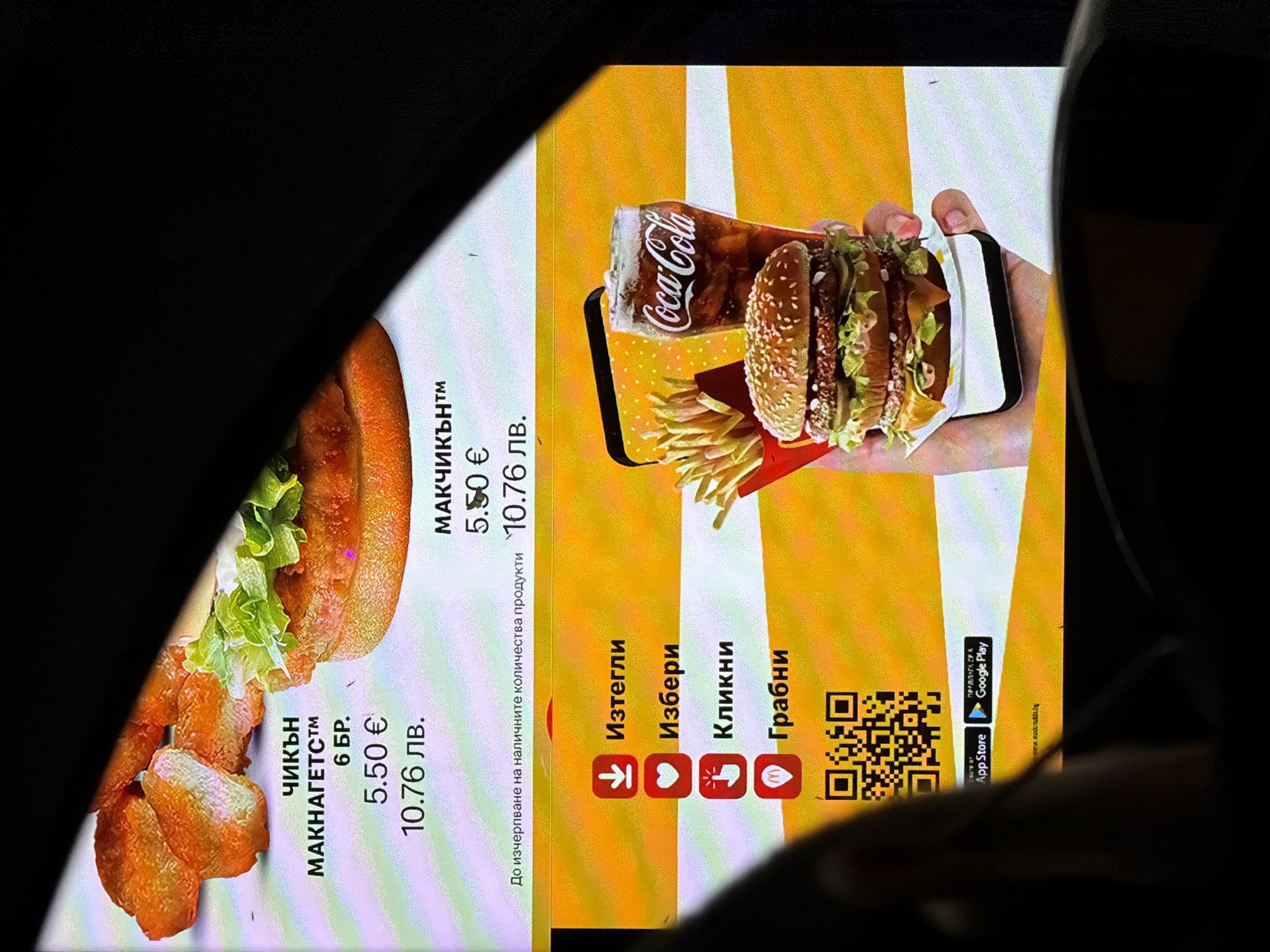 A digital menu from a fast-food restaurant, likely McDonald's, is prominently displayed, viewed from the interior of a car at what appears to be a drive-thru. The illuminated screen contrasts with the dark surroundings, suggesting it's evening or night.
The menu advertises several items. On the left, an image of chicken nuggets with lettuce and sauce is shown, accompanied by the text "ЧИКЪН МАКНАГЕТС™ 6 БР." (Chicken McNuggets™ 6 pcs.), priced at "5.50 €" (Euro) and "10.76 лв." (Bulgarian Lev). On the right, a burger, fries, and a Coca-Cola are depicted as if on a smartphone screen, held by a hand. Above this, "МАКЧИКЪН™" (McChicken™) is listed, also priced at "5.50 €" and "10.76 лв.". A disclaimer "До изчерпване на наличните количества продукти" (Until stocks last) is visible between the two product sections.
Below these offerings, a section with app-related calls to action is present: "Изтегли" (Download) with a down arrow icon, "Избери" (Select) with a heart icon, "Кликни" (Click) with a spark icon, and "Грабни" (Grab) with a stylized 'M' logo, likely representing McDonald's. A QR code is displayed for scanning. At the bottom, the "App Store" and "Google Play" logos are visible, introduced by "ПРЕДЛАГА СЕ В" (Available on), along with a partially visible URL, "m.mcdonalds.bg".
The presence of prices in both Euro and Bulgarian Lev, combined with the context of Burgas, Bulgaria, confirms the geographical setting. The media file captures a moment of consumer interaction with digital ordering systems at a fast-food establishment.