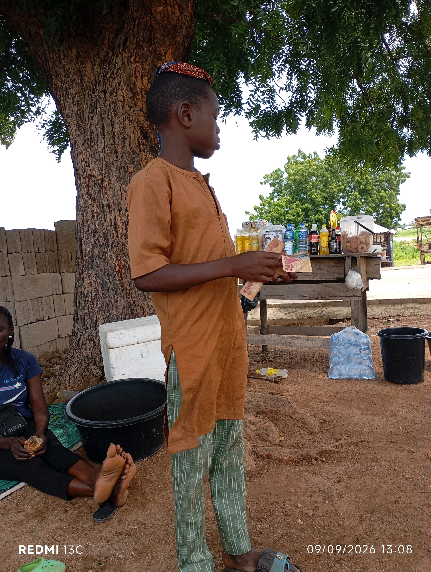 A young boy stands in a dusty, outdoor environment in Shendam, Nigeria, holding Nigerian Naira currency. He wears a brown traditional tunic and green checkered trousers, with a patterned cap on his head. To his left, a woman sits on a mat, her feet bare and resting on the ground, wearing a blue t-shirt and black pants. In the background, a wooden stall displays various bottled drinks and packaged goods, including what appear to be donuts. A large black basin and a stack of bottled water are visible on the ground. The scene is set against the backdrop of a large tree and a wall constructed of concrete blocks. The time stamp on the image indicates it was taken on September 9, 2026, at 13:08.