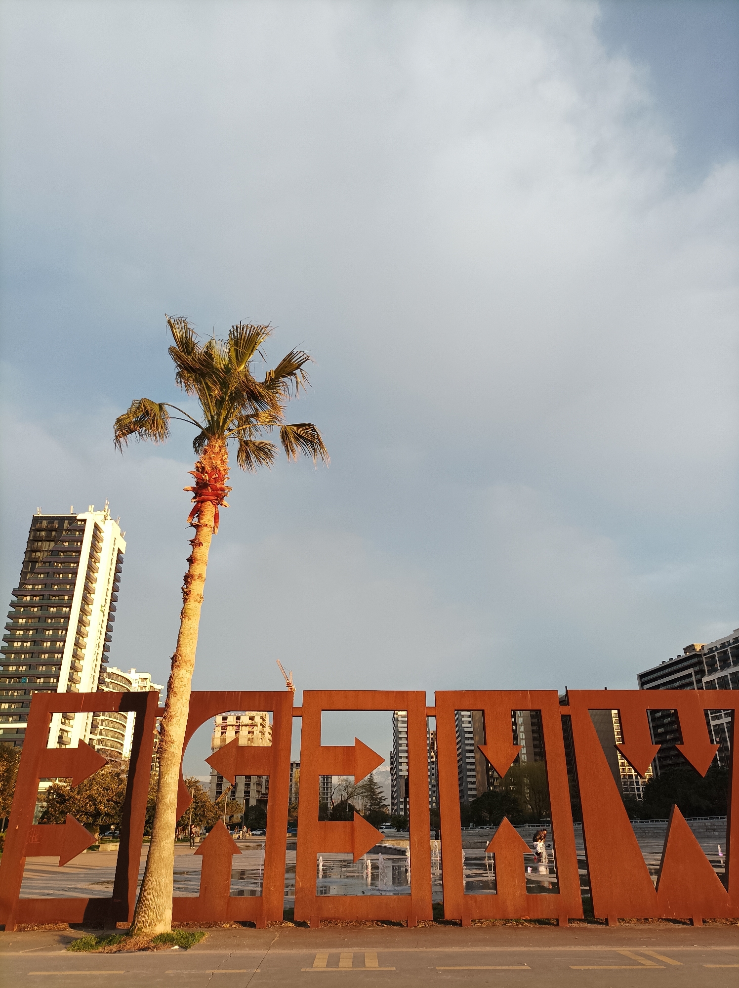 The image depicts an urban landscape in Batumi, Georgia, captured during late afternoon or early evening light. A prominent, weathered reddish-brown metal sculpture, likely Corten steel, spans the lower and mid-ground, featuring large, arrow-shaped cutouts forming letters that appear to spell "BATUMI".
In the immediate foreground, a light-colored paved surface, possibly asphalt, with faint yellow markings, occupies the bottom of the frame. A tall, slender palm tree with a light brown trunk and green fronds stands directly behind the sculpture's left-center section, its base anchored in a small patch of grass beside the pavement.
Through the cutouts of the metal sculpture, a public park or promenade area is visible. This area includes a paved walkway, green spaces, and a active fountain with multiple vertical water jets situated to the right of the palm tree. Indistinct human figures are present in this mid-ground area; one individual appears to be walking near the fountain, possibly with a stroller, while other figures are further back.
The background consists of several modern high-rise buildings. To the left, a multi-story building with a white facade and dark-paneled sections with numerous balconies is prominent. Other residential or commercial structures are visible through and above the sculpture, some showing signs of ongoing construction, including a visible construction crane. The sky is partly cloudy, with lighter patches indicating the sun's position, casting a warm, golden hue on the palm tree, the sculpture, and the illuminated facades of some buildings. Shadows are elongated, consistent with the time of day.