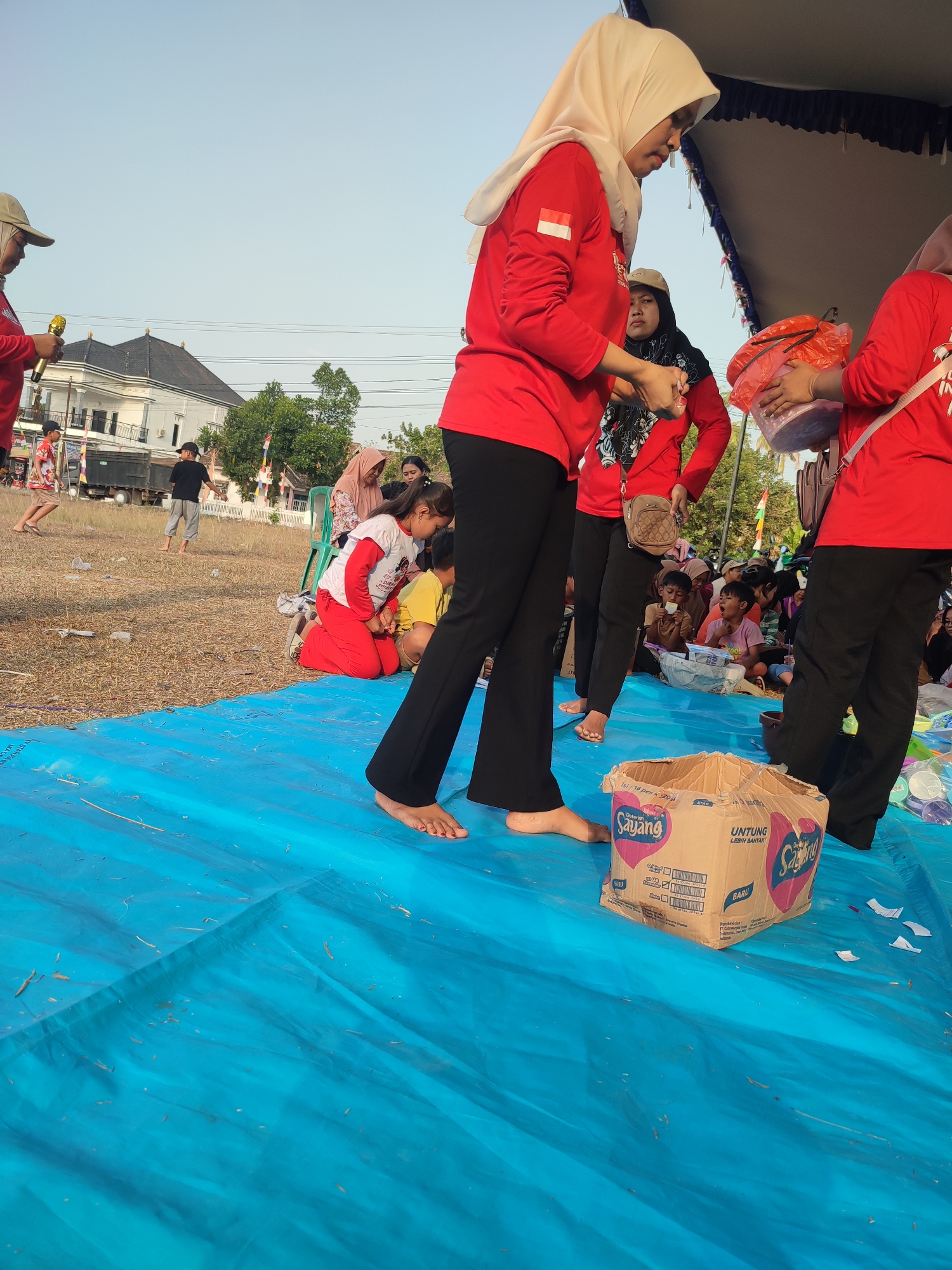 In Nusa Jaya, Indonesia, a group of people are gathered outdoors on a dry, grassy field under a clear sky. A large blue tarp covers the foreground of the scene, where a partially opened cardboard box is prominently placed. Several individuals, mostly women wearing red shirts, are visible. One woman in the foreground, wearing a cream-colored hijab and a red long-sleeved shirt with a small Indonesian flag on the sleeve, is holding a microphone. To her right, another woman in a red shirt is holding a clear plastic bag filled with items. In the background, several children and adults are seated or kneeling, watching the activities. A white building with a dark roof and trees are visible behind the gathering, suggesting a community event or festival. The ground is covered with dry grass and some scattered debris.