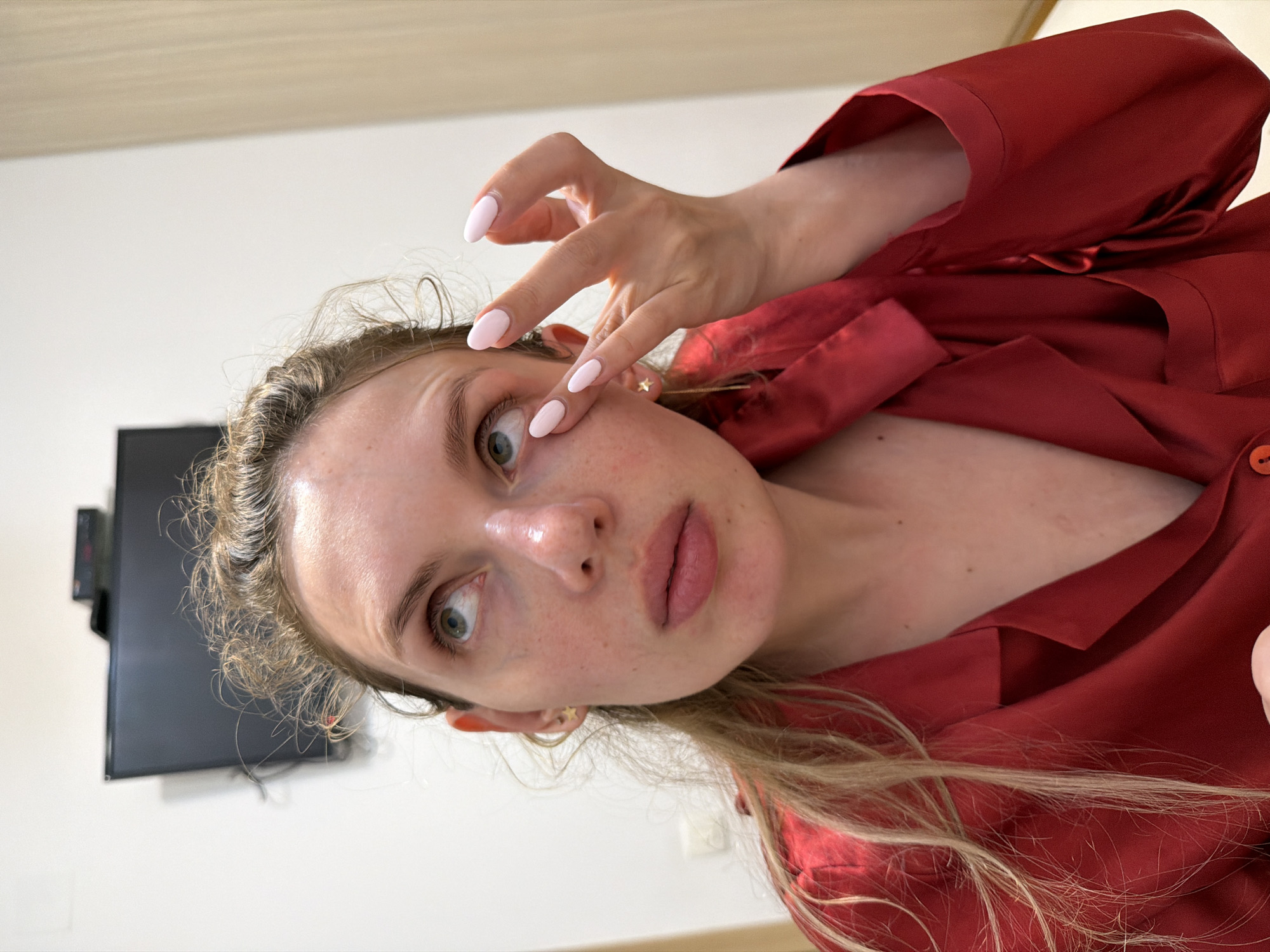 A woman with fair skin and blonde hair is shown in a close-up selfie, holding her fingers near her right eye. She is wearing a red satin shirt or pajamas. Her fingernails are painted a light pink color. In the background is a white wall, and a black television screen is partially visible. She wears a star earring on her left ear. The location is inside an apartment or room, possibly in the city of Budva, Montenegro, although there's no definitive visual cue to confirm the specific location.