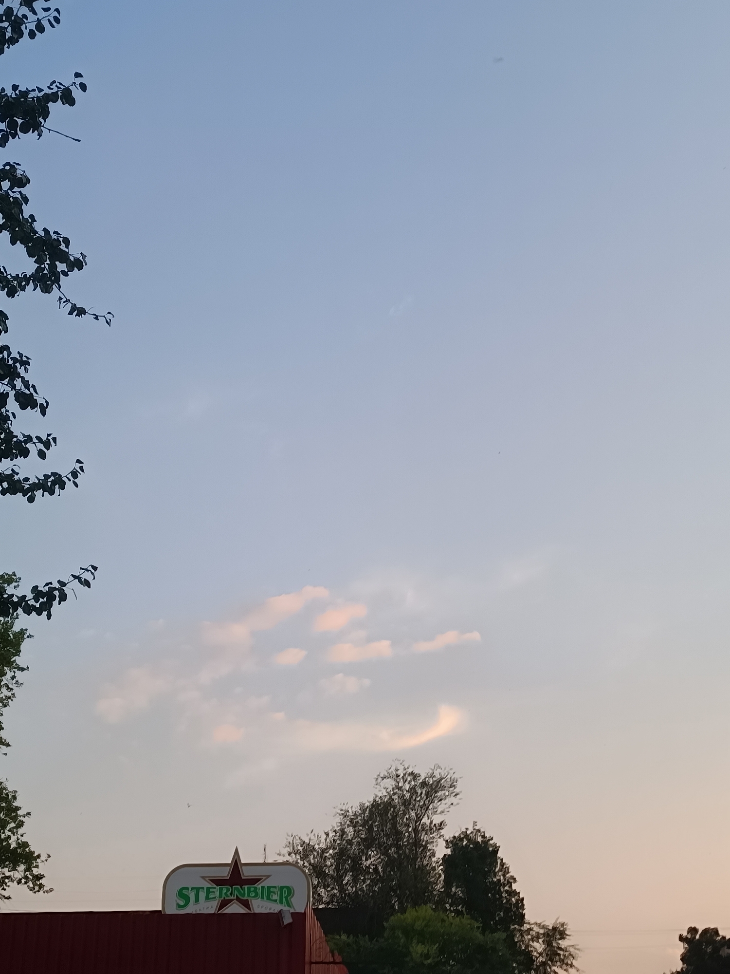 The image shows a clear sky with a few scattered clouds. In the lower part of the frame, a sign for "Sternbier" is visible, illuminated by soft light. Trees and foliage surround the sign, suggesting an outdoor setting, possibly near a building or establishment. The overall atmosphere is serene and peaceful, with the sky transitioning from a lighter hue at the horizon to a deeper blue overhead. The presence of the "Sternbier" sign indicates a commercial or recreational location.