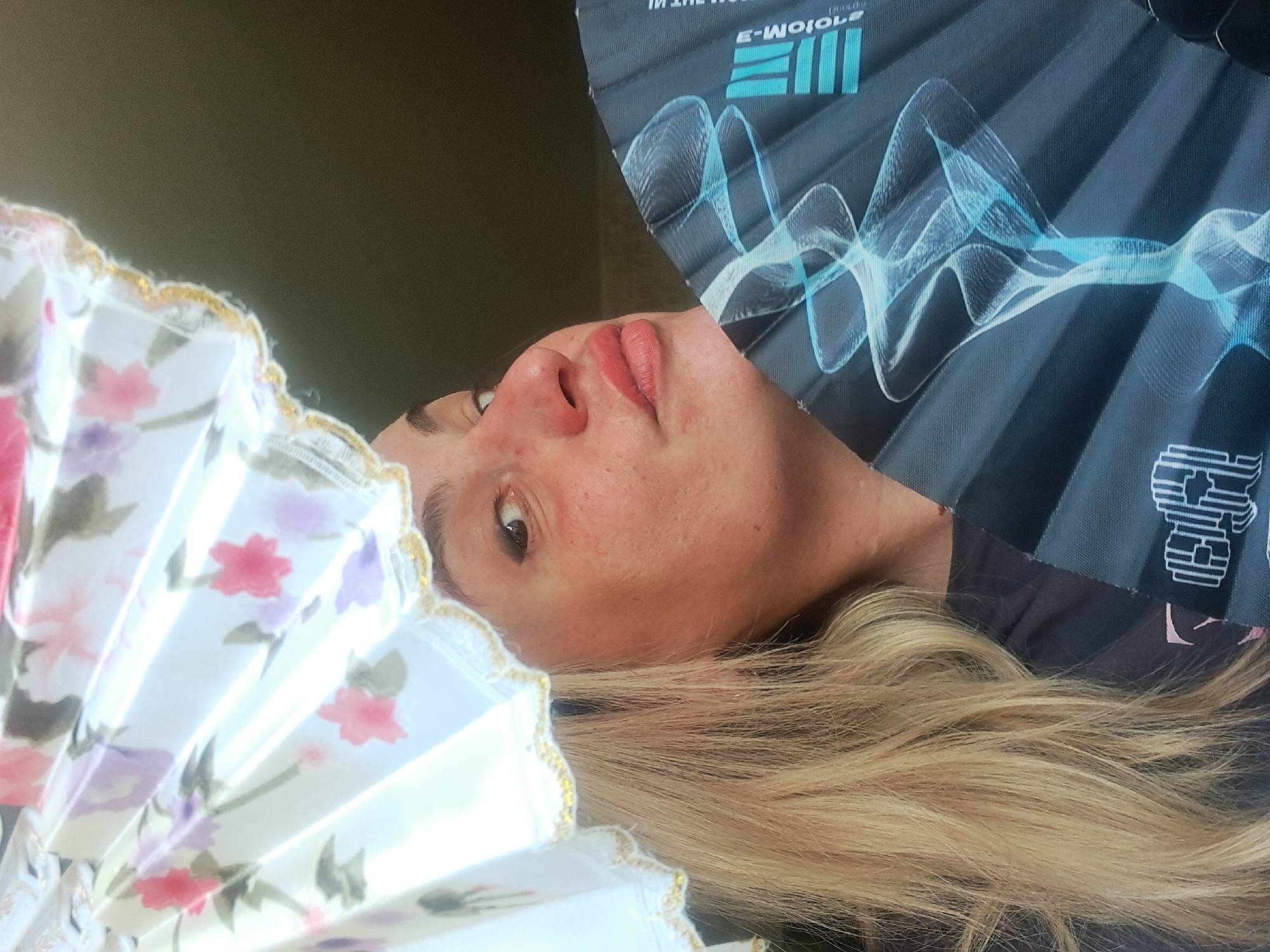 A selfie shot shows a woman in an undetailed, indoor environment. She is holding two fans, one floral-patterned in the lower left and another printed with the "E-MOJOLE" logo in the upper right. She has blonde hair and fair skin. The location is unknown.