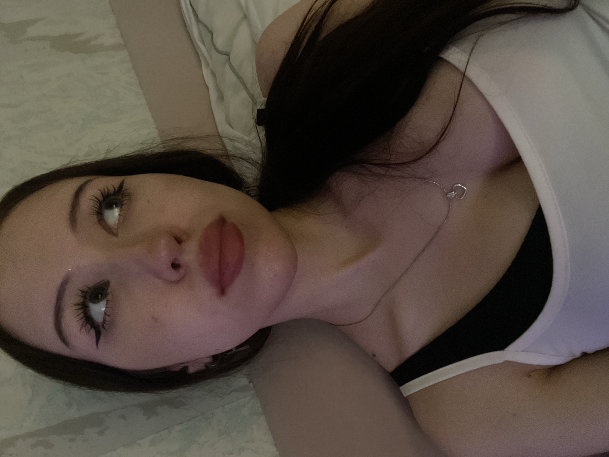 The image is a close-up, indoor selfie of a young woman lying down. She has long, dark hair pulled back into a ponytail, and is wearing a white tank top over a black sports bra. A silver necklace with a heart-shaped pendant is visible. The background appears to be a bed with light-colored bedding and a textured wall. The lighting is dim, creating a soft, diffused glow. The woman is looking off to the side with her eyes open, and her makeup includes winged eyeliner. There are no discernible elements in the image that would allow for an identification of its location within Votkinsk, Russia.