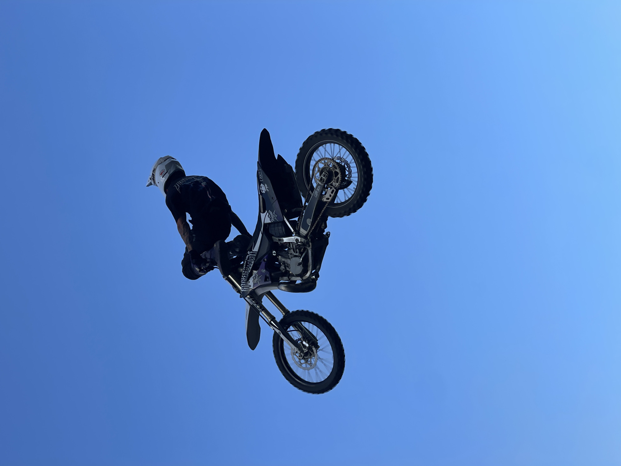 A motocross rider performs a mid-air trick against a clear blue sky. The rider, wearing a white helmet with "BELL" visible, dark clothing, and gloves, is positioned on a black and purple dirt bike. The bike is angled, with the front wheel slightly higher than the rear. The rider's body is contorted as if maneuvering the bike. The background is an uninterrupted expanse of blue sky, suggesting an outdoor event or practice session, potentially in the vicinity of Strathroy-Caradoc, Ontario, Canada, where such extreme sports events might take place given the open spaces typical of the region. No other people or objects are visible.