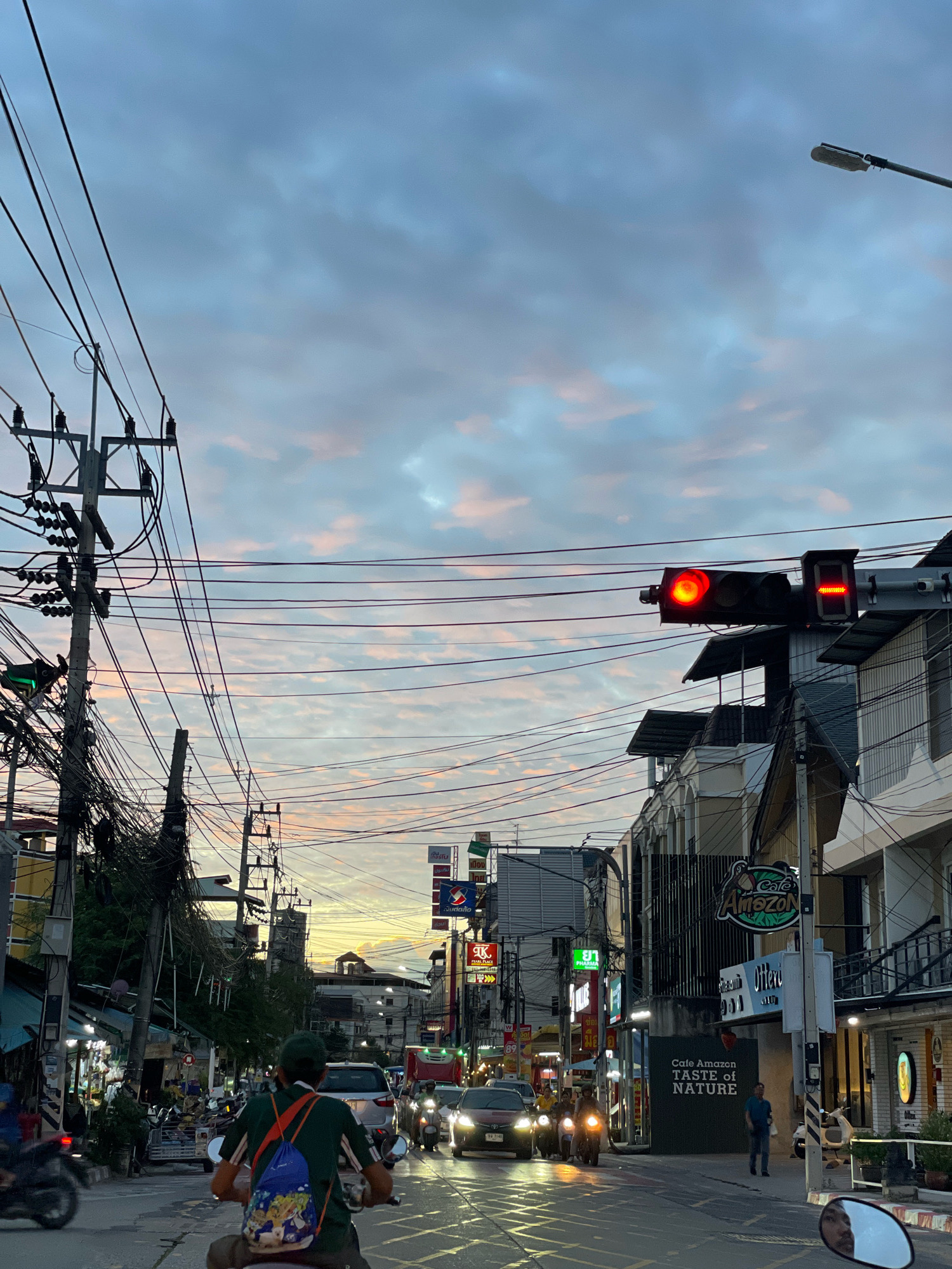 The image depicts a street scene in Pattaya City, Thailand, during twilight. The sky is filled with a mix of blue and pink clouds. A busy street is lined with buildings, power lines, and traffic signals. Several vehicles, including cars and motorcycles, are visible on the road. People are seen walking and riding on motorcycles. A traffic light is illuminated red, indicating a stop. Signs for businesses like "Cafe Amazon" are displayed on the buildings. The overall atmosphere suggests a typical evening in a bustling urban environment.