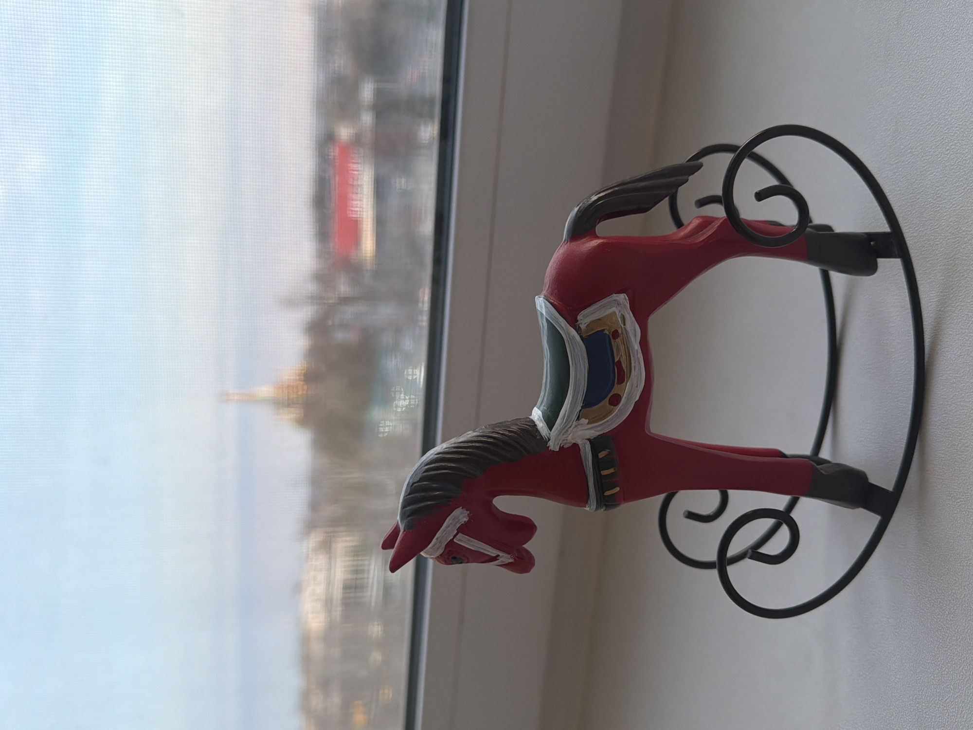 A red rocking horse with a black metal stand sits on a white windowsill. Outside the window, a blurred urban scene is visible, including the golden domes of St. Michael's Cathedral in Izhevsk, Russia.